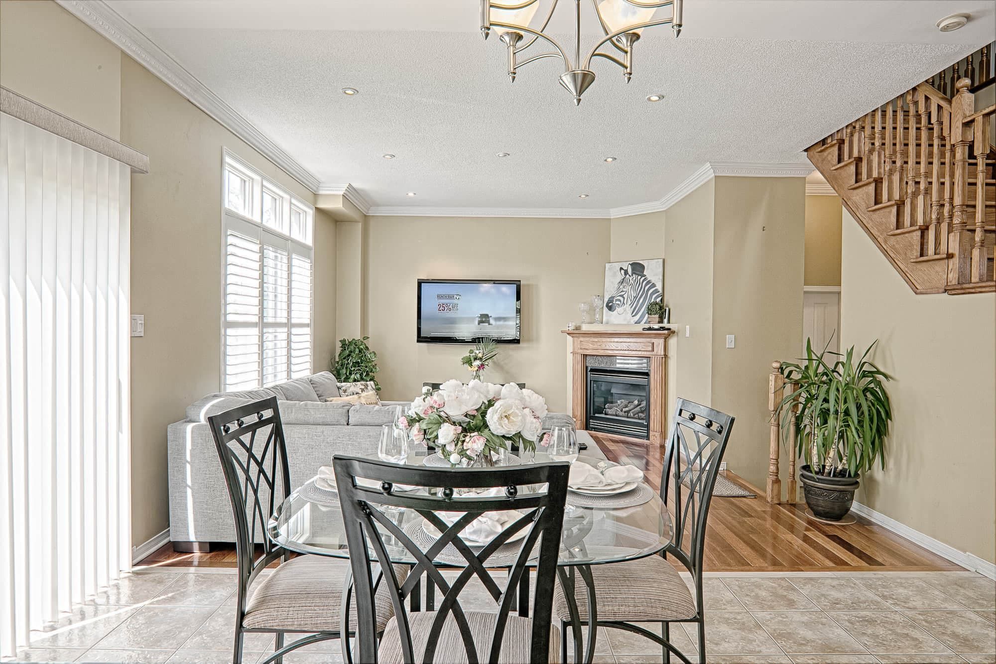 This bright traditional kitchen and dining area features rich wood cabinetry, granite countertops, and a cozy dining setup that creates a welcoming family environment. Large windows with plantation shutters allow natural light to illuminate the space, while the elegant chandelier and glass dining table enhance the overall warmth and sophistication. A beautiful example of functional kitchen renovation and classic home interior design inspiration for homeowners in Toronto and the GTA.