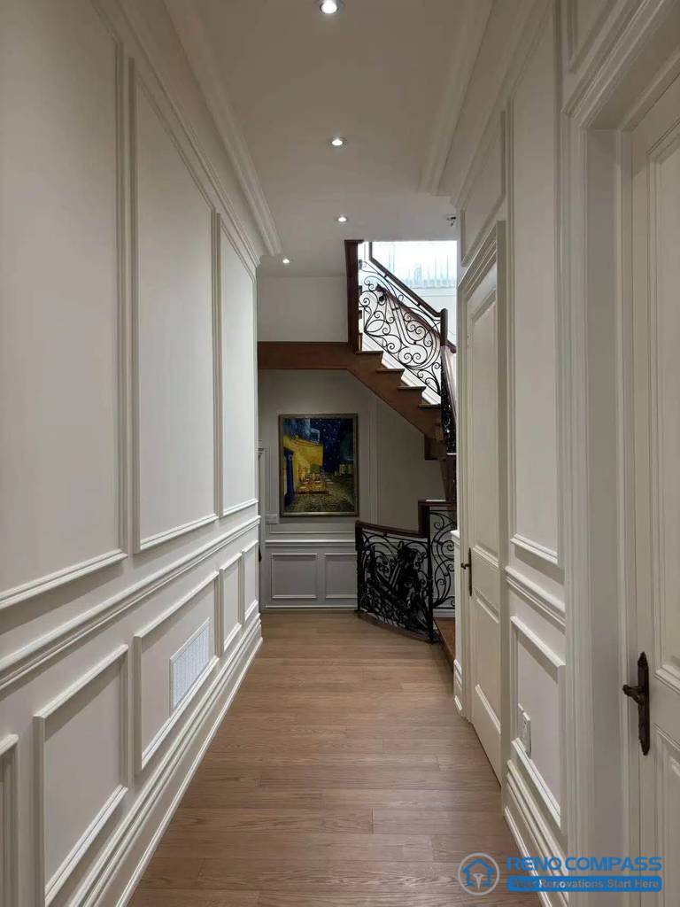 This image features a European-style hallway defined by classical wall panelling, balanced proportions, and a clear circulation axis leading toward the staircase. Full-height panels and consistent trim detailing establish rhythm and order along the corridor, while recessed lighting supports a calm and structured passage. The transition toward the staircase introduces wrought iron detailing and layered depth, reinforcing architectural continuity between levels. Hardwood flooring grounds the space and aligns visually with adjacent rooms. This view highlights disciplined spatial planning and refined detailing within a high-end Richmond Hill interior renovation.