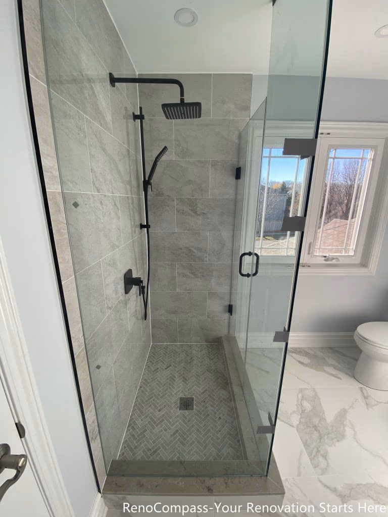 This Thornhill bathroom renovation features a walk-in glass shower with a herringbone-patterned tile base and a centered floor drain for efficient water management. Large-format gray wall tiles create a clean and structured appearance, while the frameless glass enclosure maintains visual openness and brightness. Matte black fixtures complement the neutral tones, resulting in a practical, well-proportioned shower design focused on durability, comfort, and everyday functionality.
