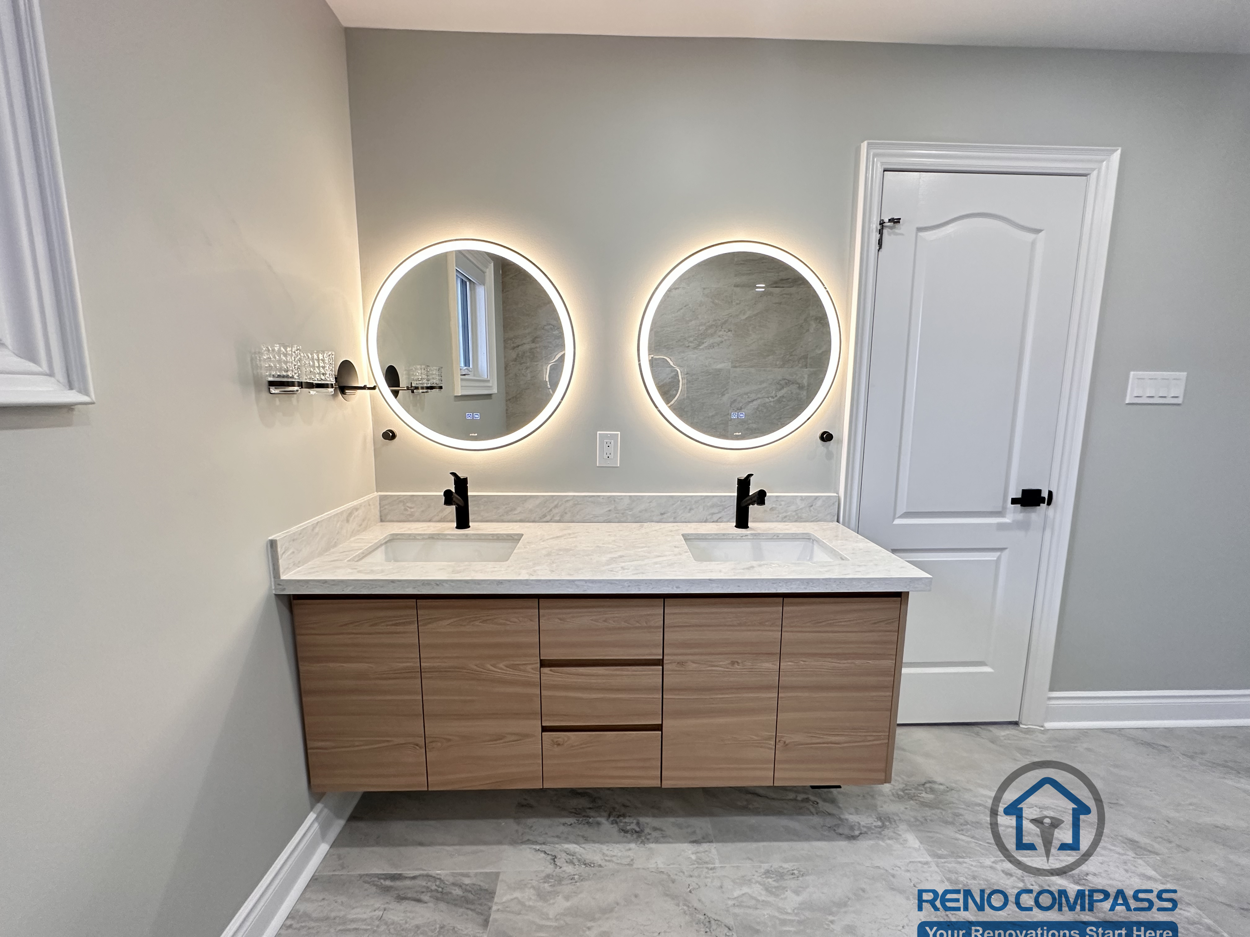 This Markham bathroom renovation showcases a floating wood-tone vanity with a quartz countertop, round LED backlit mirror, and matte black fixtures. The layout integrates a glass-enclosed shower and bathtub combination, maintaining openness and functionality. Designed and built by a professional Toronto renovation company, Reno Compass Inc., the space emphasizes clean lines, efficient circulation, and long-term usability.