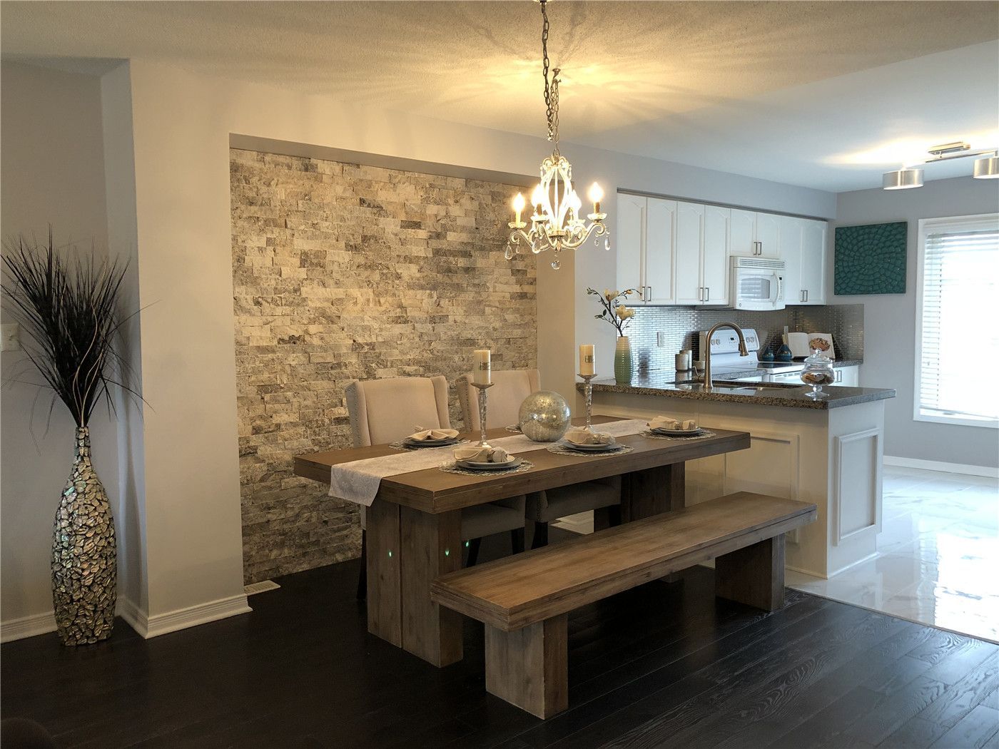 This Toronto interior design pre-sale staging showcases a warm dining area featuring a rustic wood dining table, elegant chandelier, and textured stone accent wall. The open-concept layout seamlessly connects the dining space to a bright modern kitchen with white cabinetry and sleek finishes.