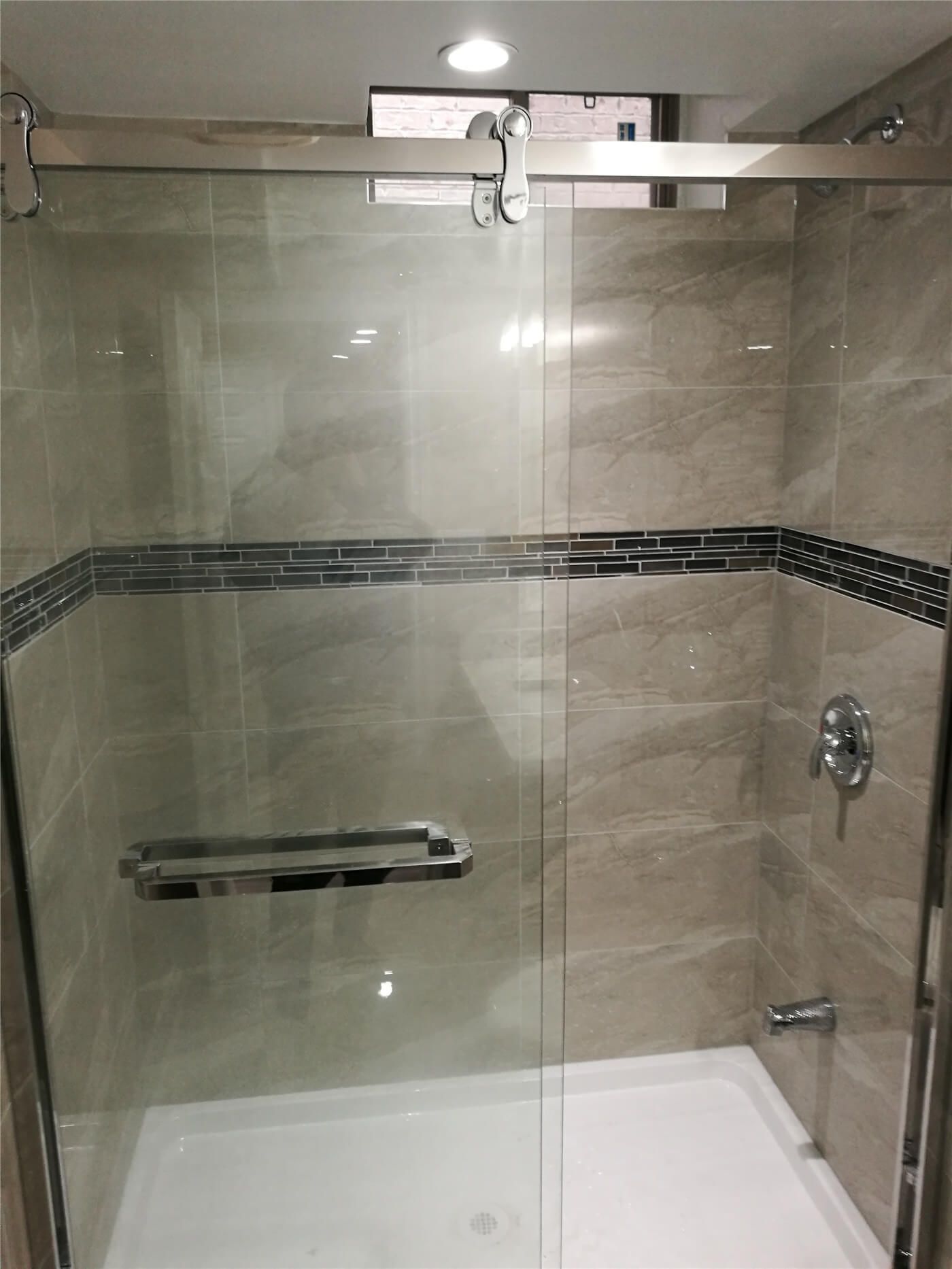 This image features a modern bathroom shower area from a Newmarket interior renovation project. The space includes a frameless glass shower enclosure, large-format wall tiles, and a decorative tile accent band that adds visual interest while maintaining a clean, contemporary look. The materials are selected for durability, waterproof performance, and easy maintenance. Completed by a professional Toronto renovation company, this bathroom renovation enhances comfort, functionality, and overall interior value.