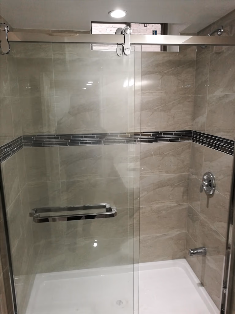 This image features a modern bathroom shower area from a Newmarket interior renovation project. The space includes a frameless glass shower enclosure, large-format wall tiles, and a decorative tile accent band that adds visual interest while maintaining a clean, contemporary look. The materials are selected for durability, waterproof performance, and easy maintenance. Completed by a professional Toronto renovation company, this bathroom renovation enhances comfort, functionality, and overall interior value.