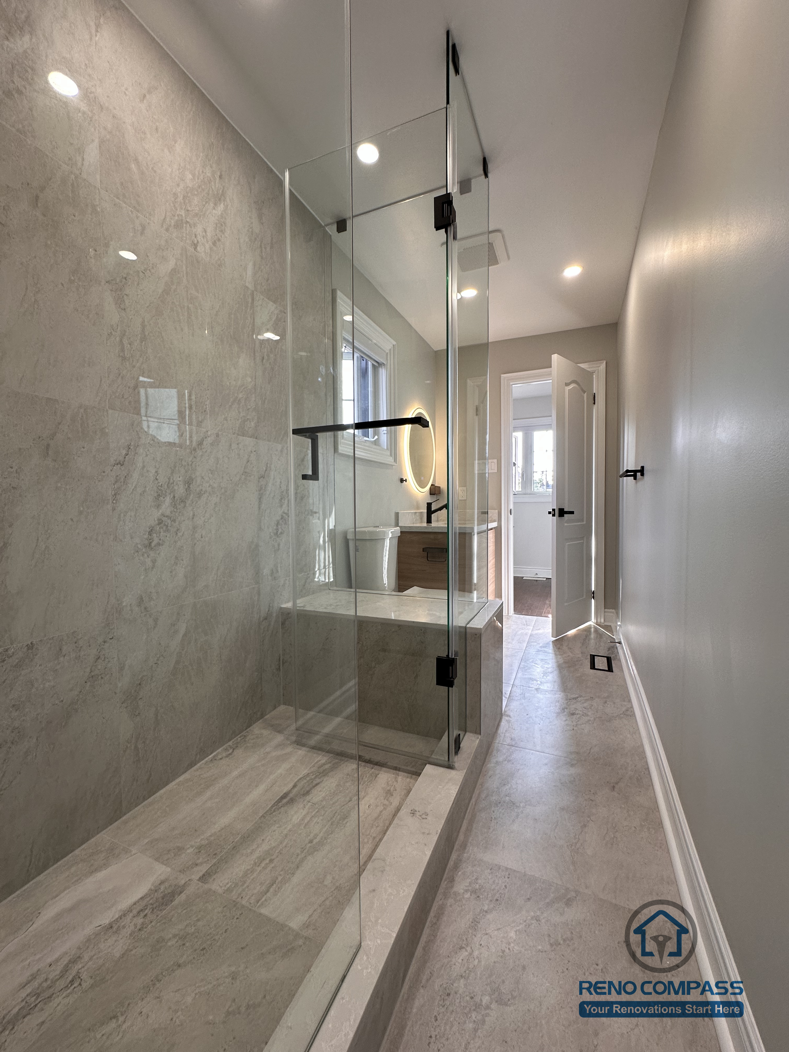 This Markham bathroom renovation features a walk-in glass shower with large-format stone-look wall and floor tiles, creating a clean and streamlined bathing area. The transparent glass enclosure maintains visual continuity along the bathroom corridor while ensuring effective water containment. Designed and built by a professional Toronto renovation company, Reno Compass Inc., the space prioritizes durability, clarity, and long-term functionality.