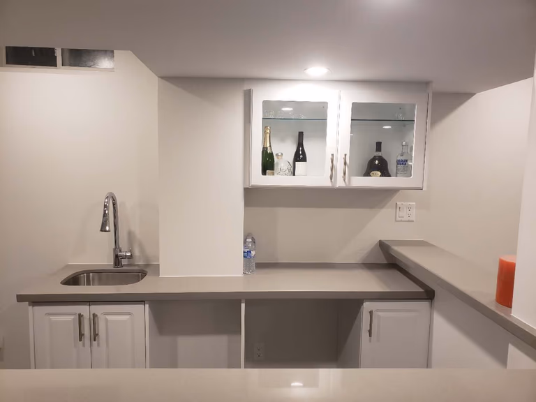 This Markham basement renovation features a clean and modern bar area with custom white cabinetry, a built-in sink, and a functional island countertop. Recessed pot lights and smooth ceiling finishes enhance brightness, while dark flooring adds contrast and warmth. The layout is designed to maximize usability and flow, making the basement ideal for entertaining, family gatherings, or multi-purpose living. This project reflects professional basement renovation standards in Markham, focusing on durability, lighting, and practical space planning.
