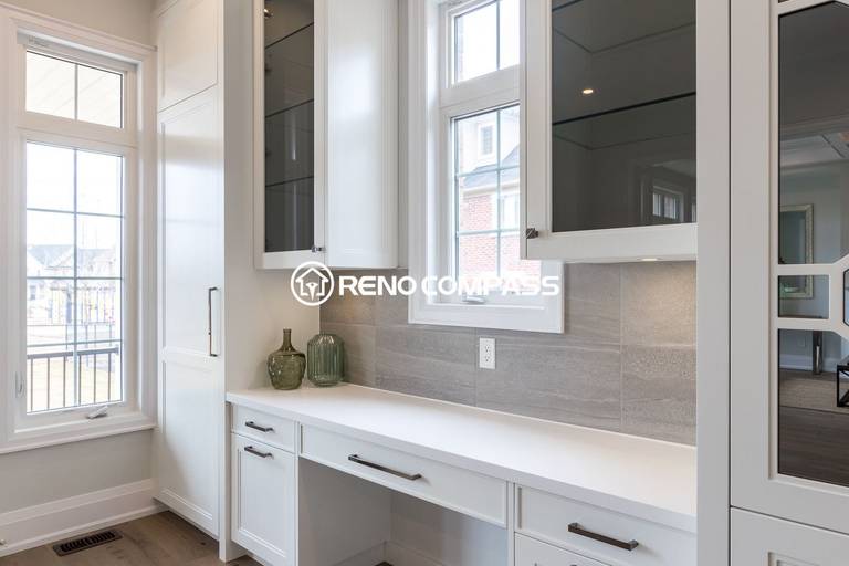 This Newmarket kitchen renovation features custom white cabinetry with glass-front storage, a quartz countertop workspace, and a modern tile backsplash. Large windows bring abundant natural light into the kitchen, enhancing the bright and elegant interior design. Completed by a trusted Toronto renovation company, Reno Compass Inc., the project highlights functional storage solutions and refined craftsmanship.