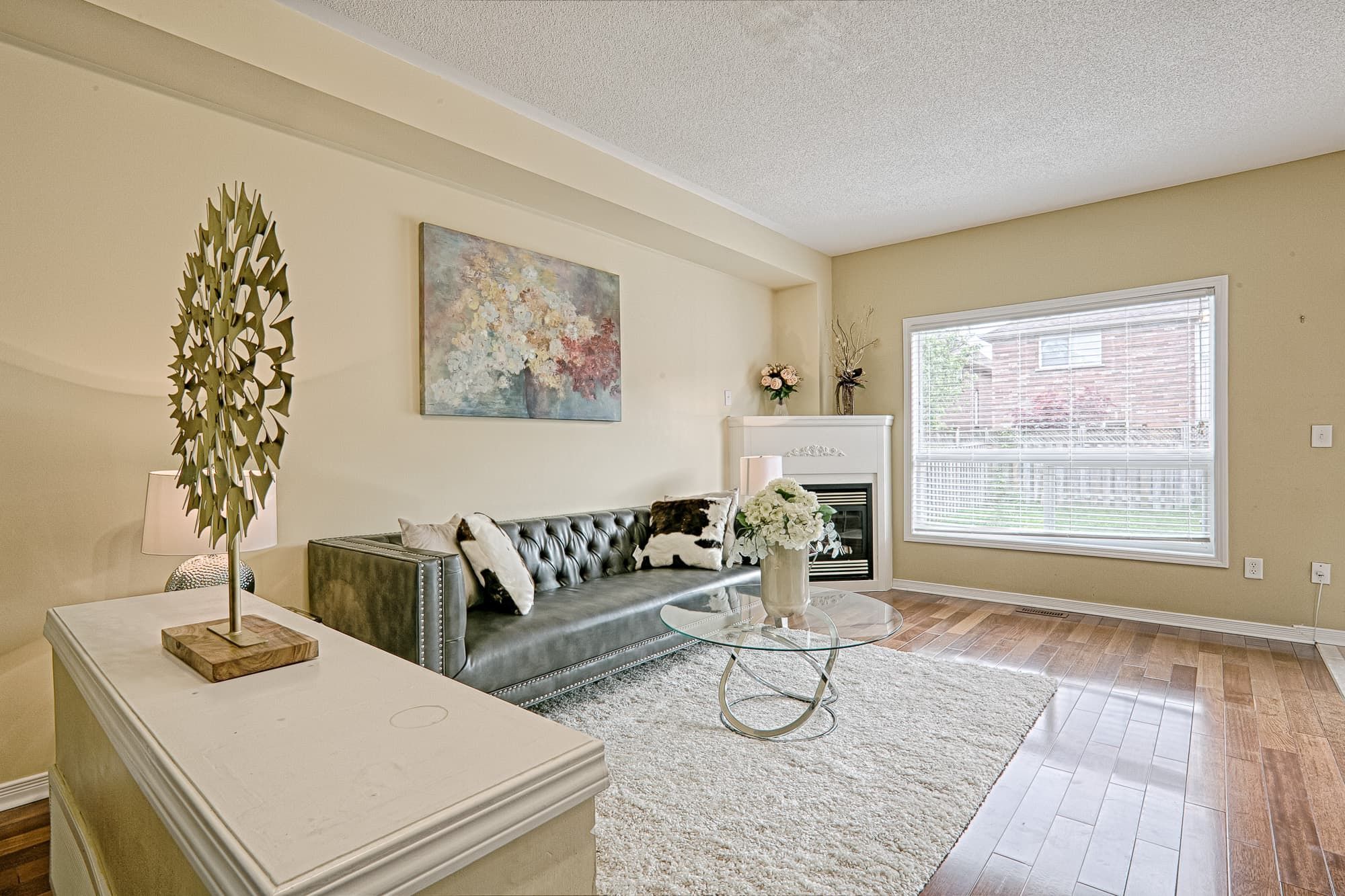 This bright living room features a simple and lovely interior design with warm hardwood flooring, a cozy fireplace, and a large window that brings in abundant natural light. The neutral color palette, soft area rug, and clean furniture layout create an inviting and comfortable living space. This design emphasizes brightness, openness, and everyday functionality, making it ideal for modern family homes.