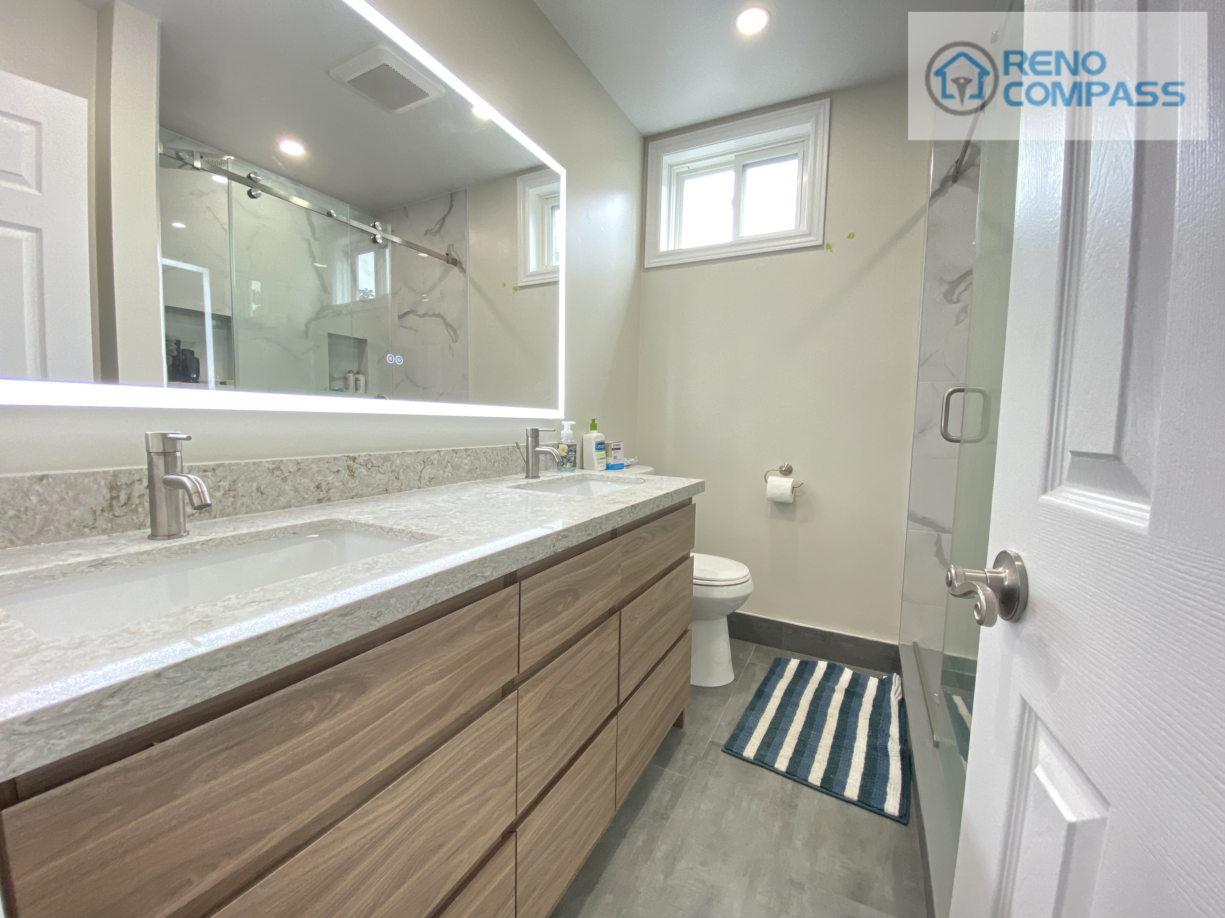 This modern bathroom renovation in Pickering features a wood-tone floating vanity with integrated storage, quartz countertop, undermount sink, and a full-size LED backlit mirror. Neutral wall tones, marble-look shower tiles, and clean-lined fixtures create a bright and functional space designed for daily use. This bathroom upgrade was completed by Reno Compass Inc. as part of a full interior renovation project.