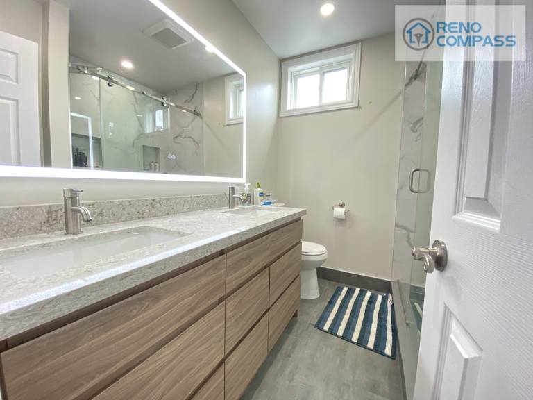This modern bathroom renovation in Pickering features a wood-tone floating vanity with integrated storage, quartz countertop, undermount sink, and a full-size LED backlit mirror. Neutral wall tones, marble-look shower tiles, and clean-lined fixtures create a bright and functional space designed for daily use. This bathroom upgrade was completed by Reno Compass Inc. as part of a full interior renovation project.