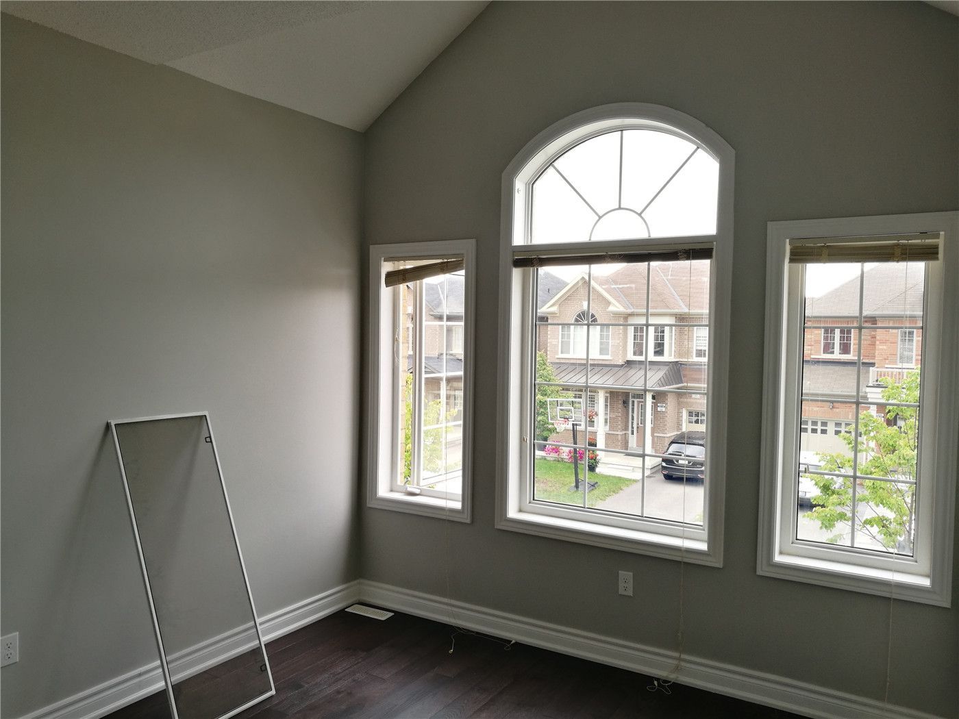 This image showcases a high-ceiling bedroom after a Markham interior renovation, featuring an arched feature window, large side windows, neutral wall tones, and dark hardwood flooring that enhances natural light and spaciousness.