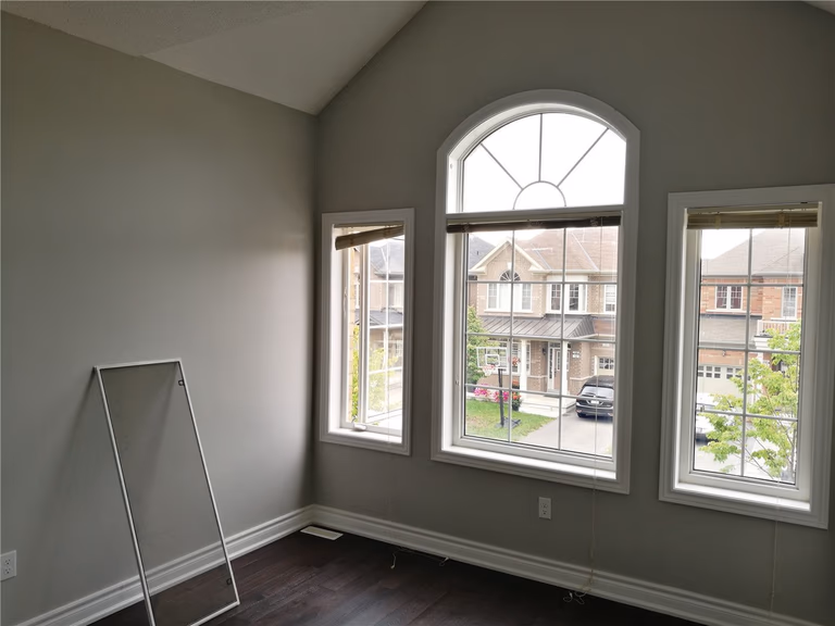 This image showcases a high-ceiling bedroom after a Markham interior renovation, featuring an arched feature window, large side windows, neutral wall tones, and dark hardwood flooring that enhances natural light and spaciousness.