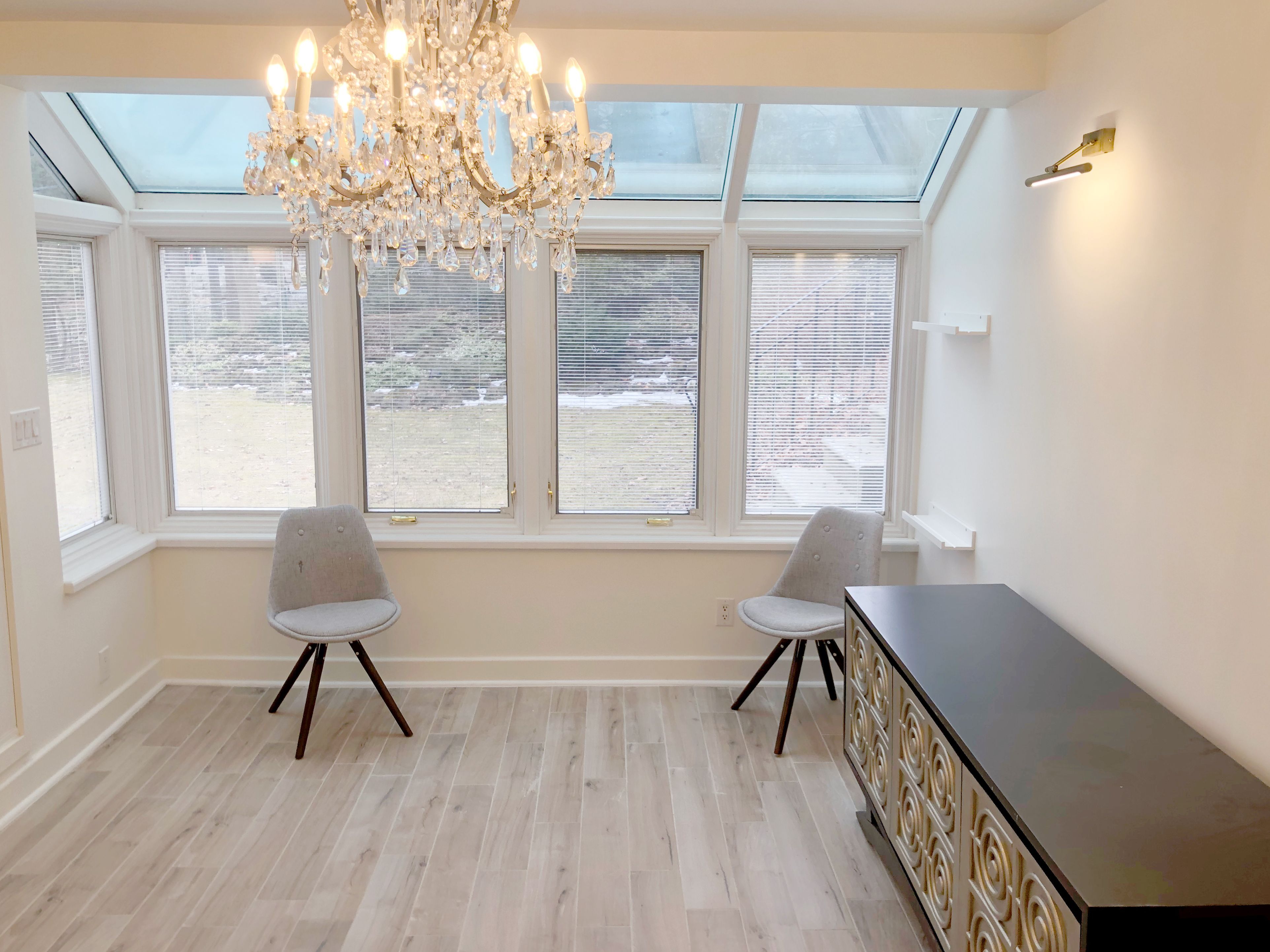 This Etobicoke interior renovation features a bright sunroom upgrade with large surrounding windows, a skylight ceiling, and an elegant crystal chandelier as the focal point. Light-toned flooring enhances the natural brightness, while clean wall finishes and minimal built-in shelving maintain a refined and airy atmosphere. Designed and completed by Reno Compass Inc., this renovation transforms the space into a comfortable and inviting sitting area that blends natural light with timeless interior design.
