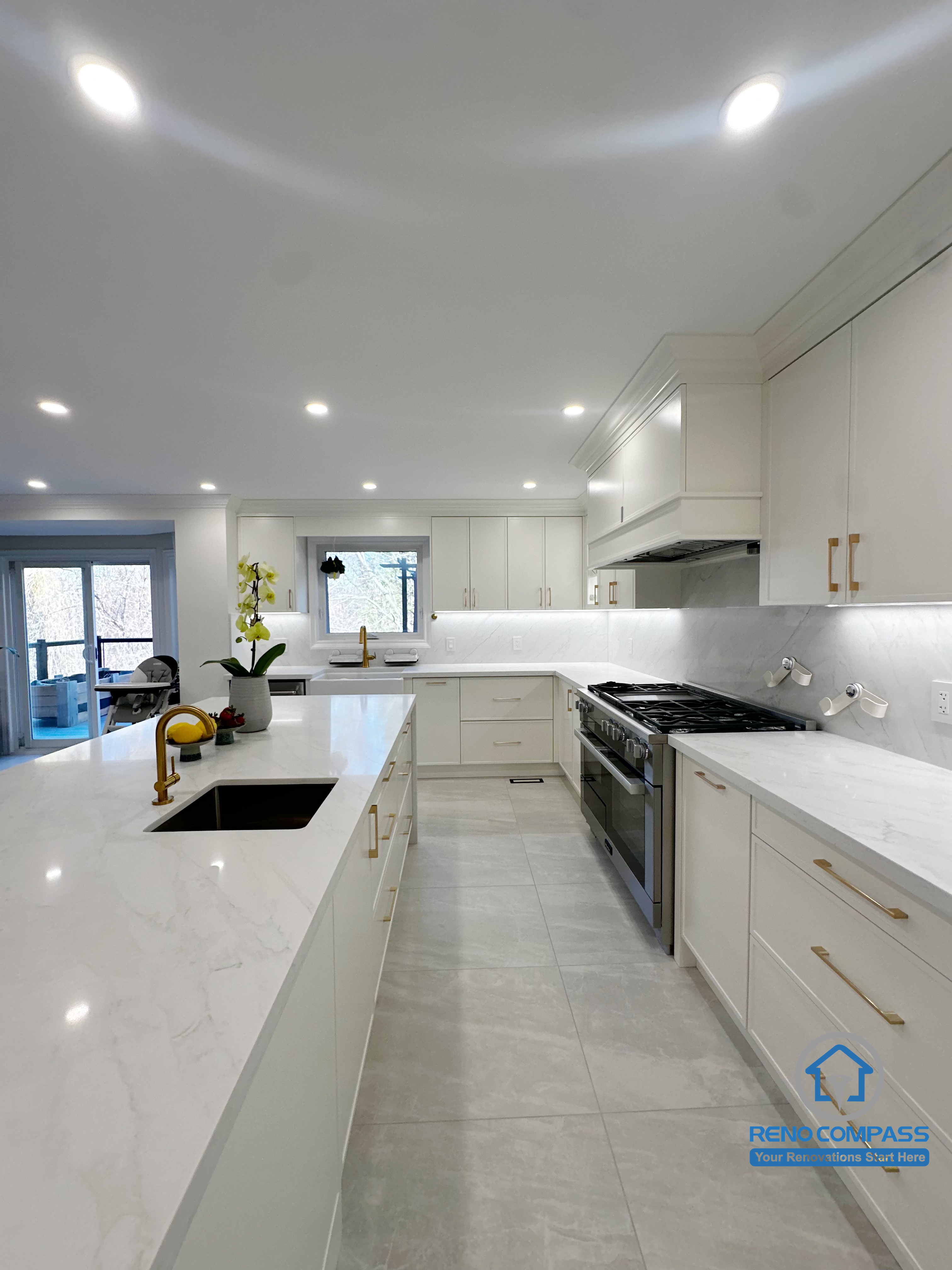 This kitchen renovation showcases an open-concept layout centered around a large island with an integrated sink, creating an efficient workflow between the sink, cooktop, and surrounding prep areas. White cabinetry, quartz countertops, and gold-toned hardware add a refined finish, while recessed lighting keeps the space bright and highly functional for everyday cooking and entertaining.
