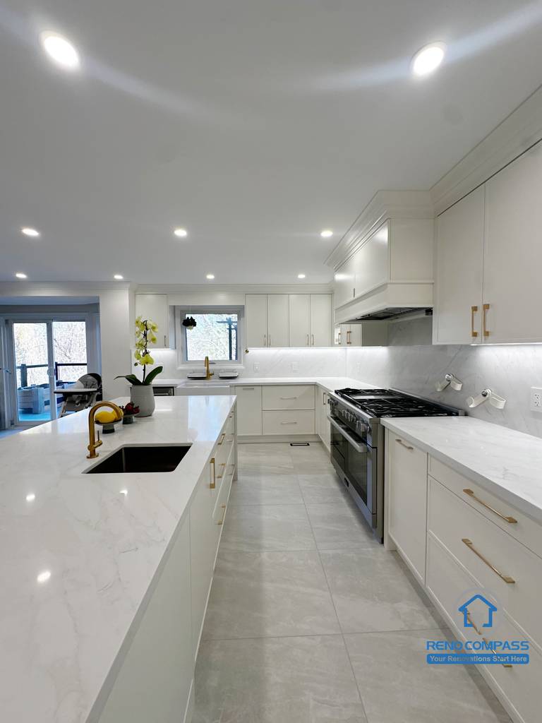 This kitchen renovation showcases an open-concept layout centered around a large island with an integrated sink, creating an efficient workflow between the sink, cooktop, and surrounding prep areas. White cabinetry, quartz countertops, and gold-toned hardware add a refined finish, while recessed lighting keeps the space bright and highly functional for everyday cooking and entertaining.