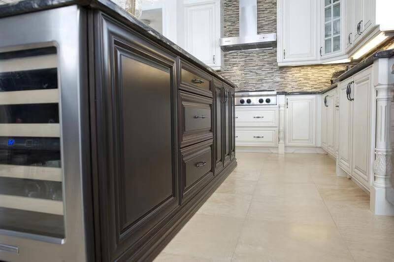 This European-style kitchen renovation showcases a custom dark wood island with detailed panel molding and integrated storage drawers, creating a bold contrast against the surrounding white cabinetry. The island features a built-in wine cooler, adding both convenience and upscale functionality to the space.

The natural stone countertop enhances durability and elegance, while the textured stone backsplash and stainless steel range hood provide a refined focal point in the background. Neutral tile flooring completes the cohesive, high-end design.

This layout highlights premium craftsmanship and thoughtful appliance integration, ideal for homeowners seeking a luxury kitchen renovation with custom millwork and functional island features.