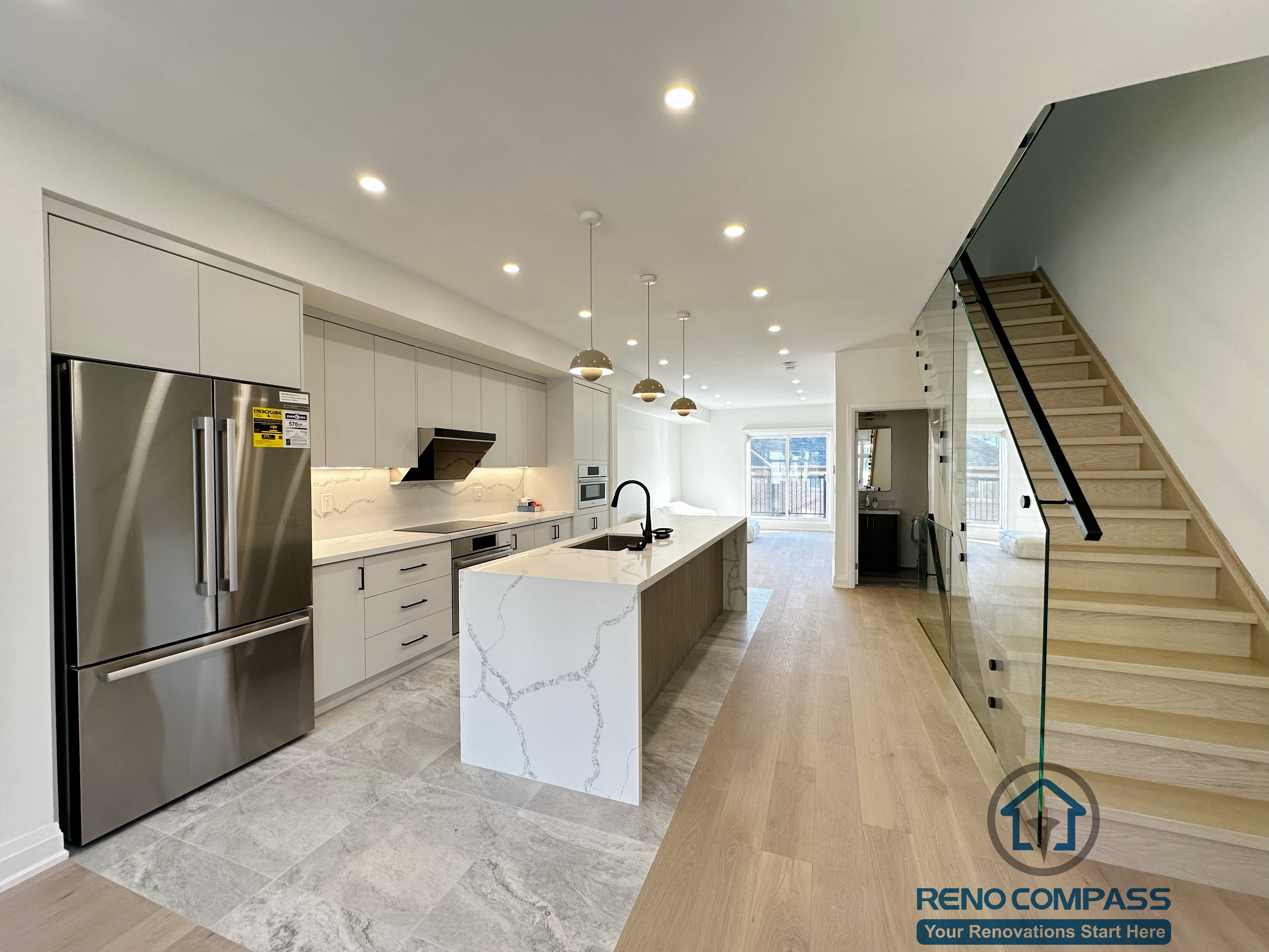 This image showcases a modern kitchen renovation in Richmond Hill, featuring a large waterfall quartz island, full-height cabinetry, stainless steel appliances, and a sleek glass staircase. The combination of tiled kitchen flooring and natural wood floors enhances the open-concept layout, creating a bright and functional main living space.