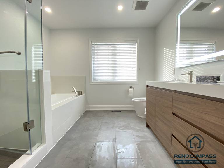 This Markham bathroom renovation features a spacious layout combining a modern bathtub, frameless glass shower, and a custom double-sink vanity with integrated LED mirror. Large-format floor tiles enhance durability and visual continuity, while natural light improves overall comfort. Completed by a trusted Toronto renovation company, this bathroom prioritizes functionality, clean design, and long-term usability.