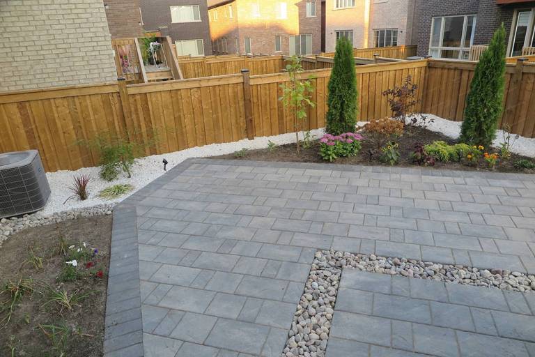 Backyard landscaping project in Vaughan featuring a modern interlocking patio with integrated decorative river rock drainage channels, garden beds, and cedar privacy fencing. The design combines functional hardscaping with clean landscape lines to create a low-maintenance and visually balanced outdoor living space.