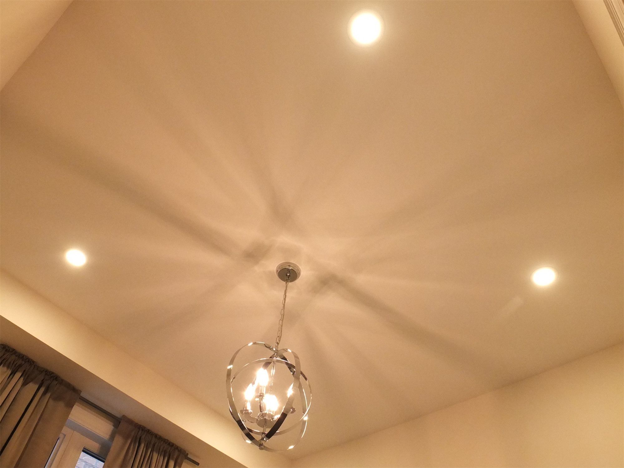 This Kleinburg interior renovation features a stylish ceiling design highlighted by a contemporary chandelier paired with recessed pot lights. The combination of ambient ceiling lighting and decorative fixture creates a warm and elegant atmosphere while enhancing the architectural lines of the room. This lighting arrangement adds both visual interest and balanced illumination to the modern living space.