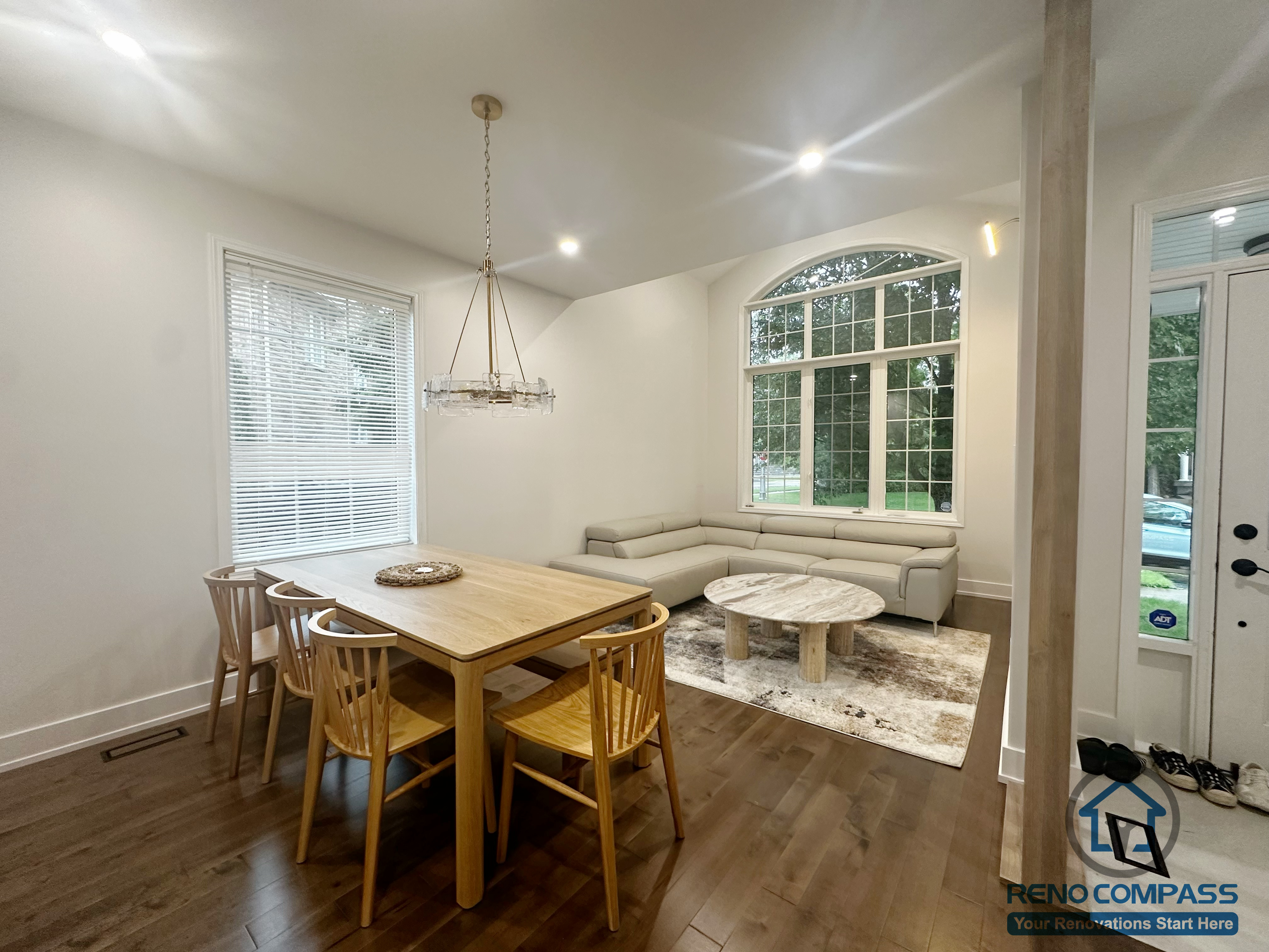 This Oakville interior renovation features an open-concept dining and living area designed to maximize natural light and everyday comfort. The dining space is anchored by a solid wood table and minimalist lighting fixture, creating a warm and functional setting for daily meals. Adjacent to the dining area, the living space is framed by a large arched window that brings in abundant daylight and enhances the sense of openness. Continuous hardwood flooring visually connects both areas, while the balanced layout supports smooth circulation and long-term livability within a modern family home.