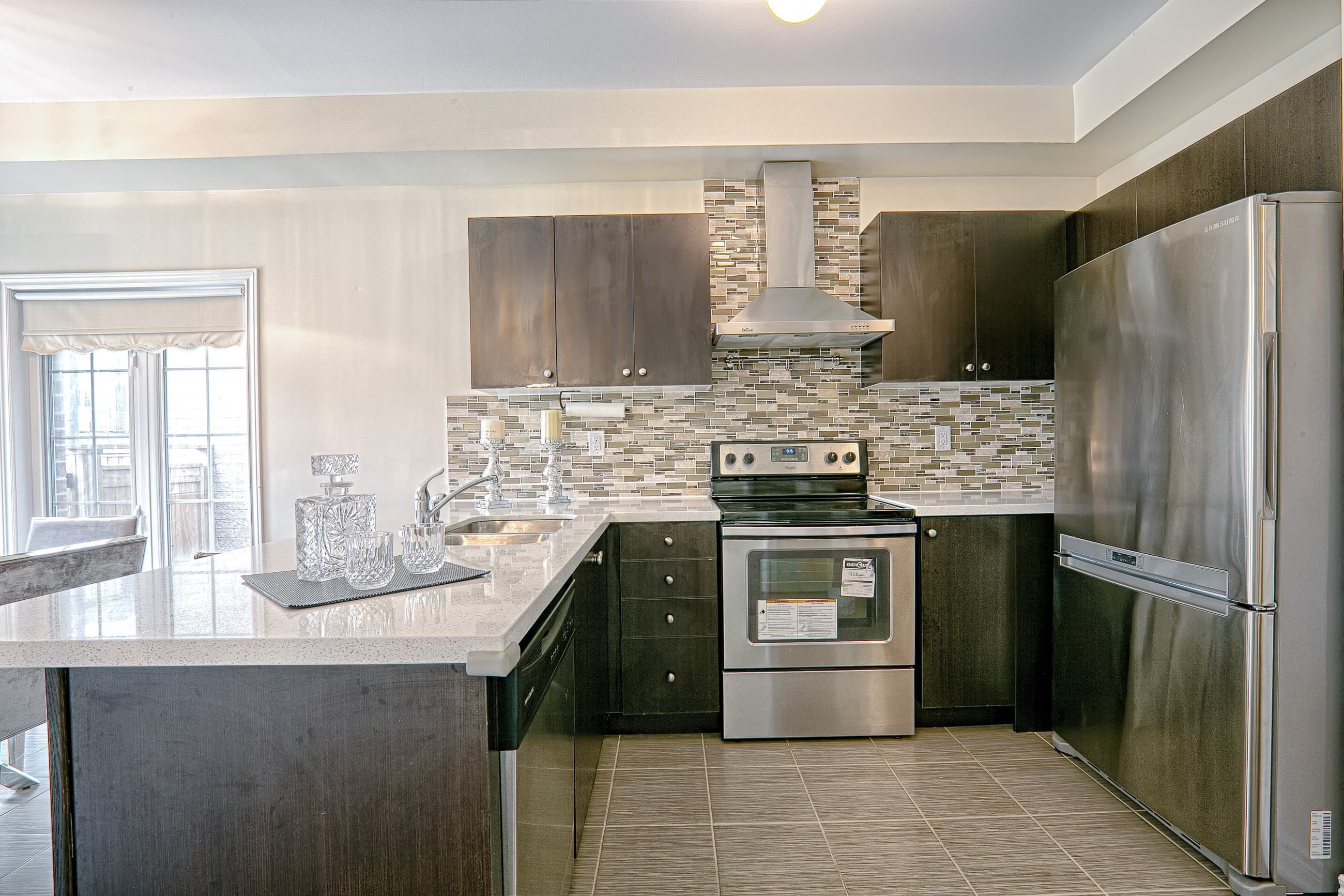 This modern L-shaped kitchen renovation showcases dark wood cabinetry, sleek stainless steel appliances, and durable quartz countertops designed for both style and practicality. The mosaic tile backsplash adds texture and visual depth, while the functional layout maximizes storage and workflow efficiency. Finished with contemporary floor tiles and a clean neutral palette, this kitchen reflects the expertise of a trusted Toronto renovation company delivering high-quality residential interior upgrades with modern design appeal.