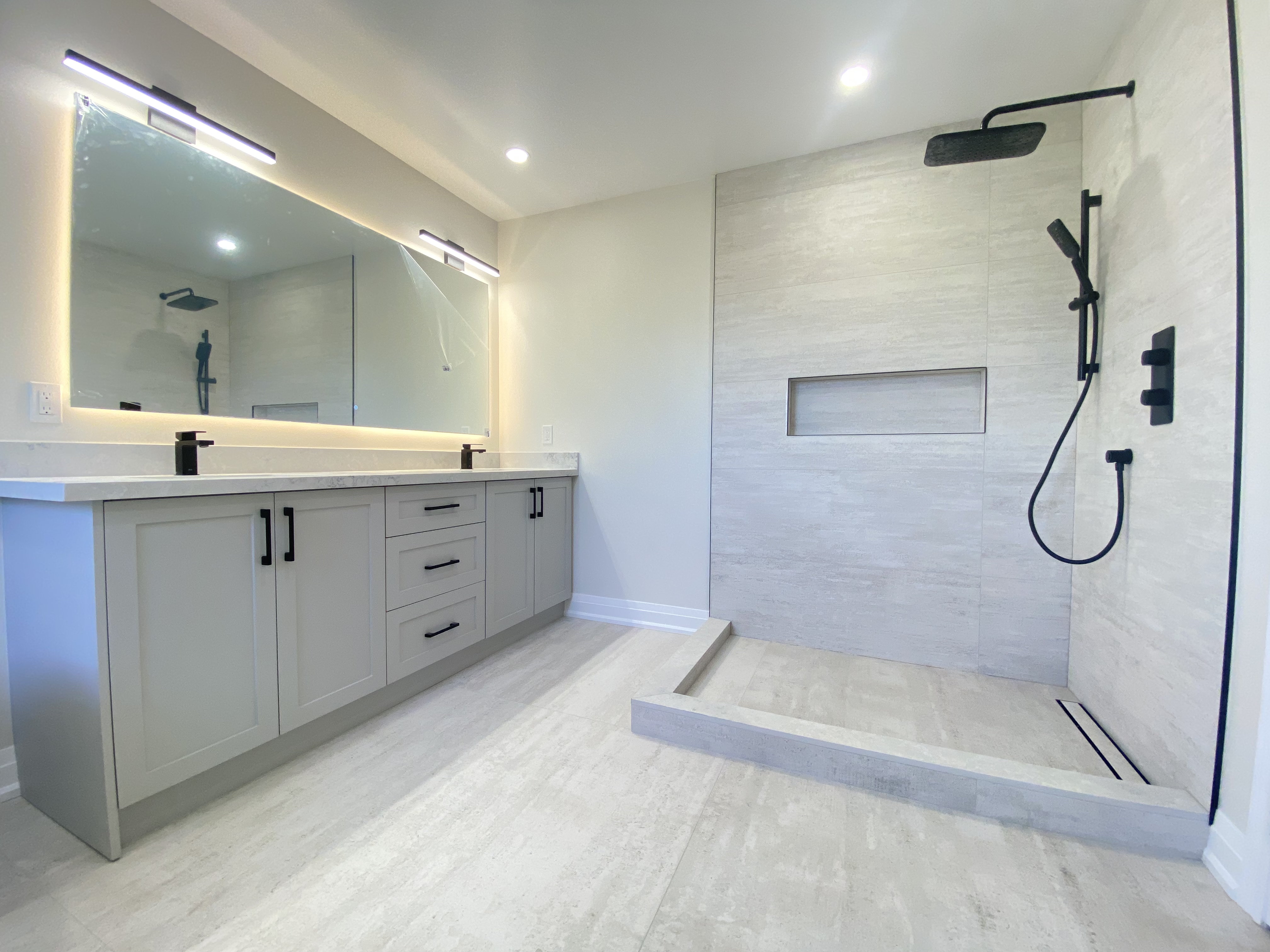 This bathroom renovation in Richmond Hill features a custom double vanity with integrated storage, LED backlit mirror lighting, and matte black fixtures paired with a modern walk-in shower. The large-format tile flooring and wall finishes create a clean, cohesive look throughout the space.