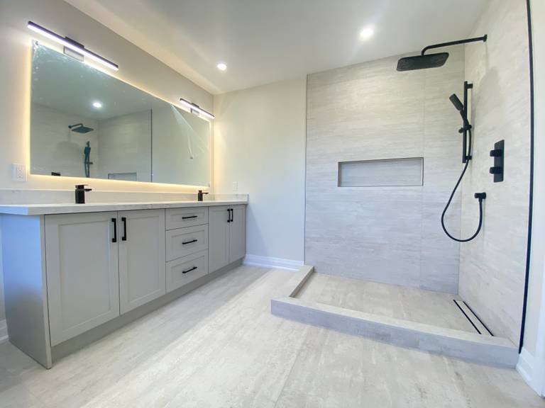This bathroom renovation in Richmond Hill features a custom double vanity with integrated storage, LED backlit mirror lighting, and matte black fixtures paired with a modern walk-in shower. The large-format tile flooring and wall finishes create a clean, cohesive look throughout the space.