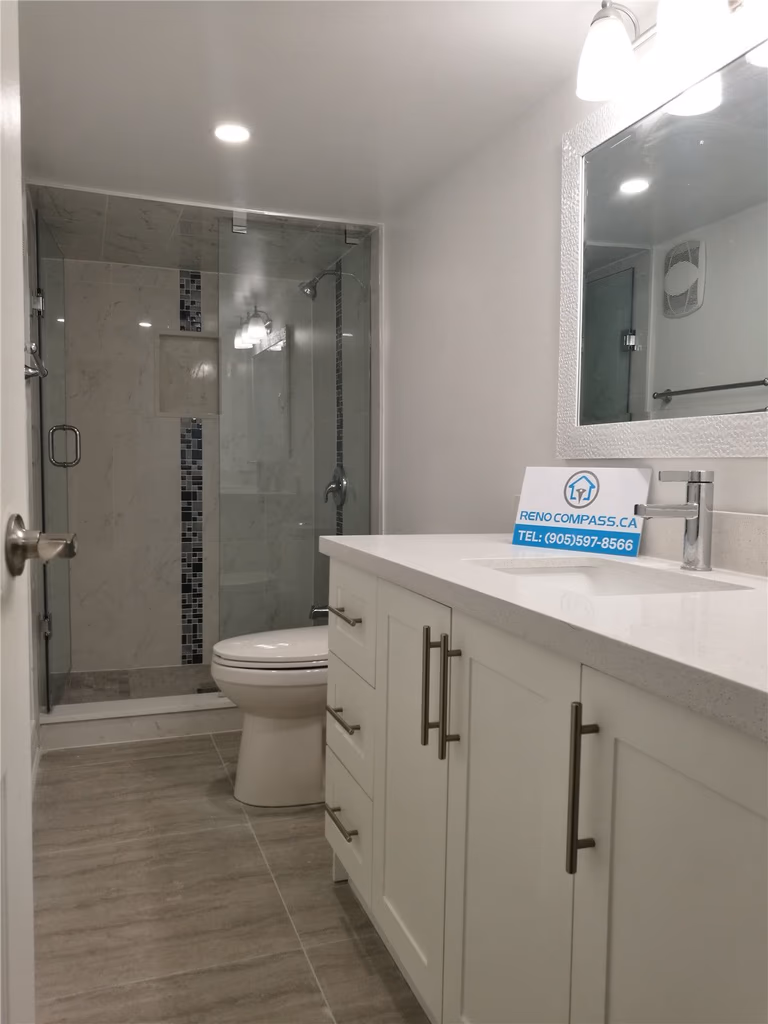 Bathroom renovation project in Pickering featuring a modern white vanity with quartz countertop, undermount sink, large mirror, and a frameless glass shower enclosure. The neutral tile flooring and clean layout enhance brightness and functionality, completed by a professional Toronto renovation company.
