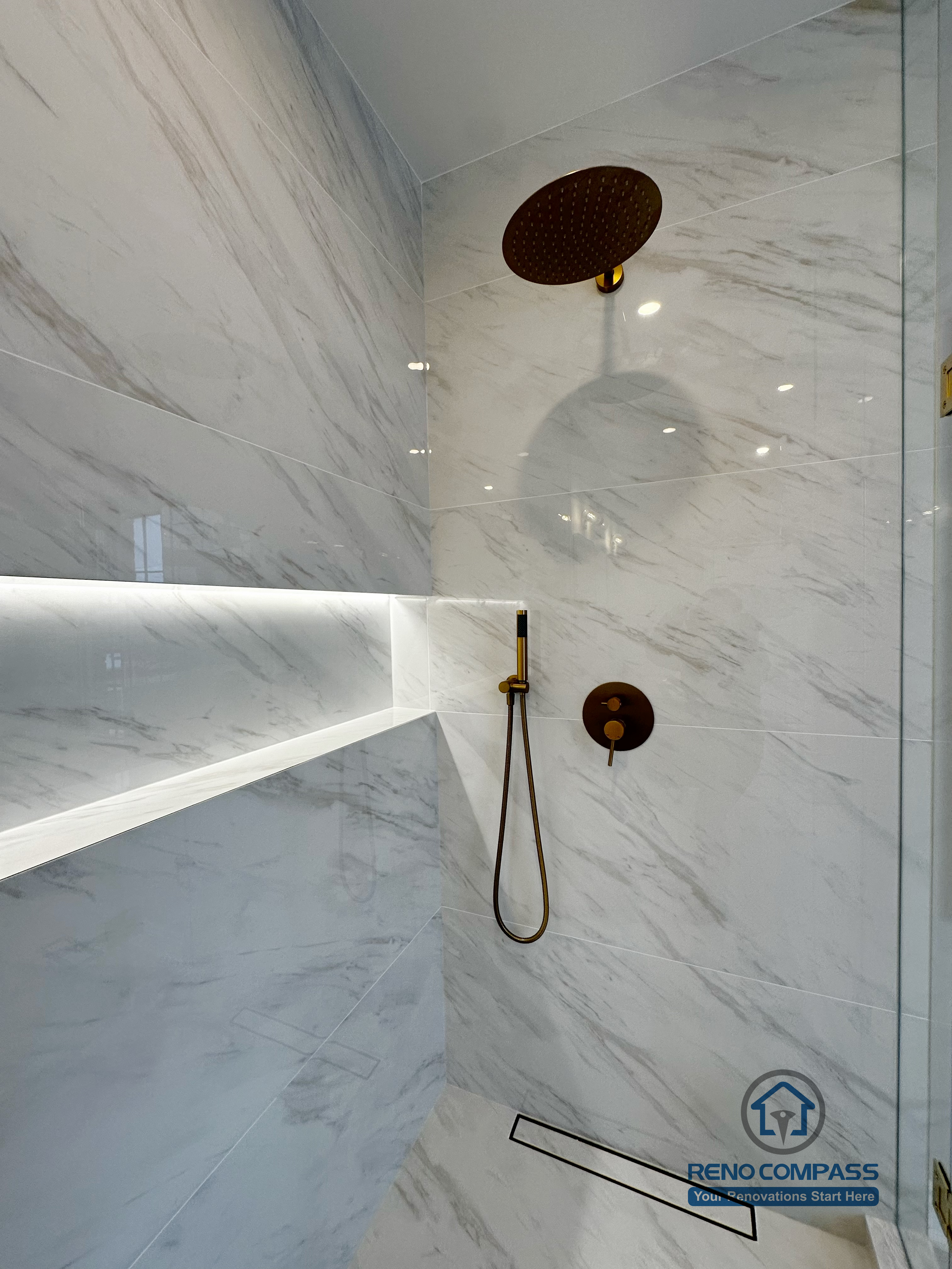 Close-up view of a custom shower detail in a Vaughan primary bathroom renovation, featuring a rainfall shower head, handheld fixture with gold hardware, marble-look porcelain tile walls, and an integrated LED-lit shower niche. This refined shower design was completed by a professional Toronto renovation company with a focus on luxury finishes, comfort, and precision installation.