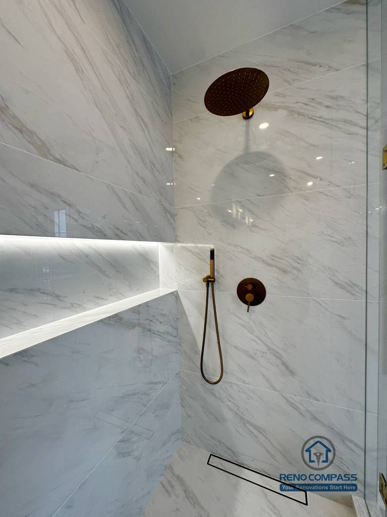 Close-up view of a custom shower detail in a Vaughan primary bathroom renovation, featuring a rainfall shower head, handheld fixture with gold hardware, marble-look porcelain tile walls, and an integrated LED-lit shower niche. This refined shower design was completed by a professional Toronto renovation company with a focus on luxury finishes, comfort, and precision installation.