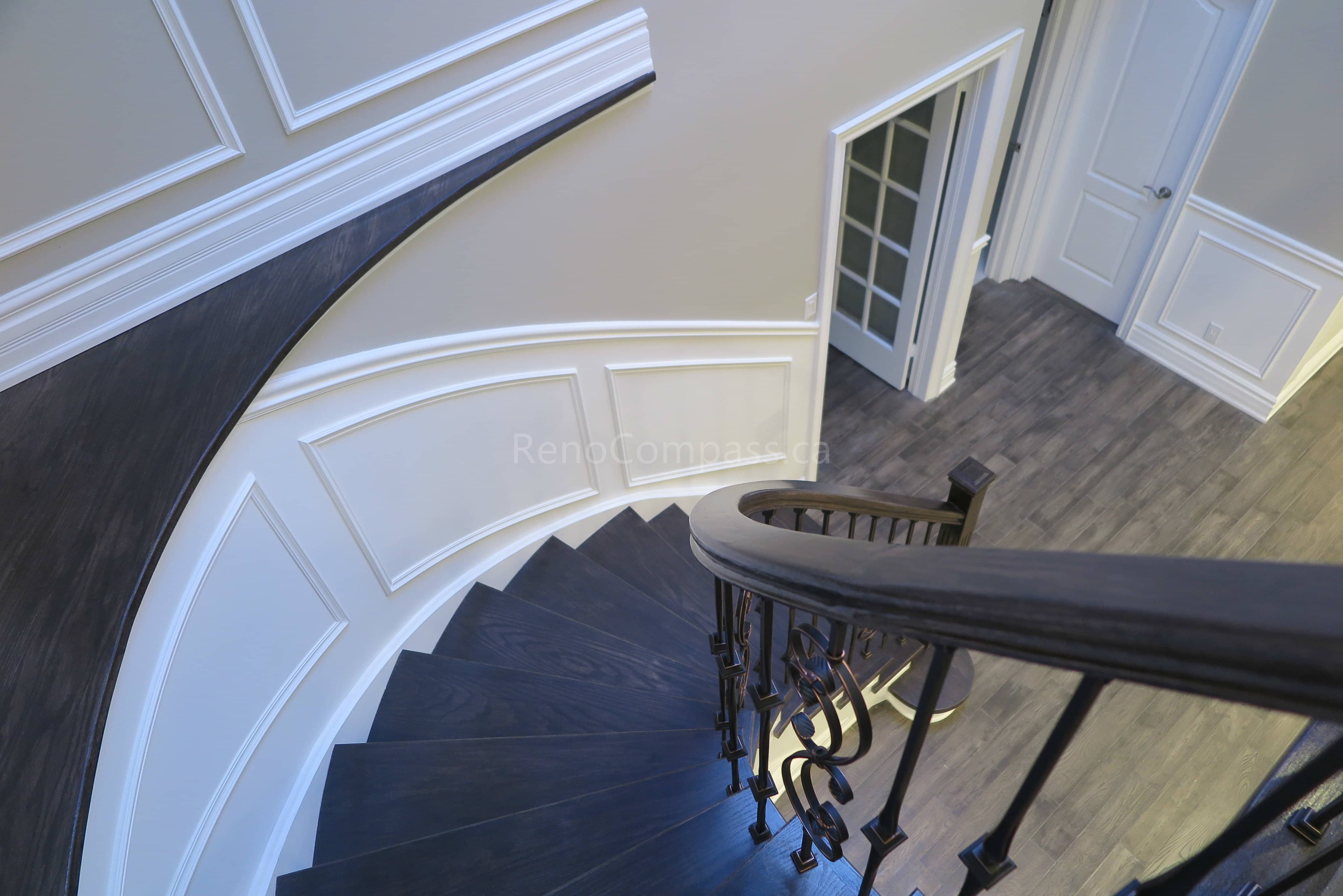 This Markham interior renovation features a stunning custom curved staircase with dark wood treads, white risers, and elegant wrought iron railings. Detailed wall panel molding and modern flooring enhance the sophisticated architectural design, creating a luxurious focal point within the home. Completed by Reno Compass, a professional renovation contractor specializing in custom staircases and high-end interior renovations across Markham and the Greater Toronto Area.
