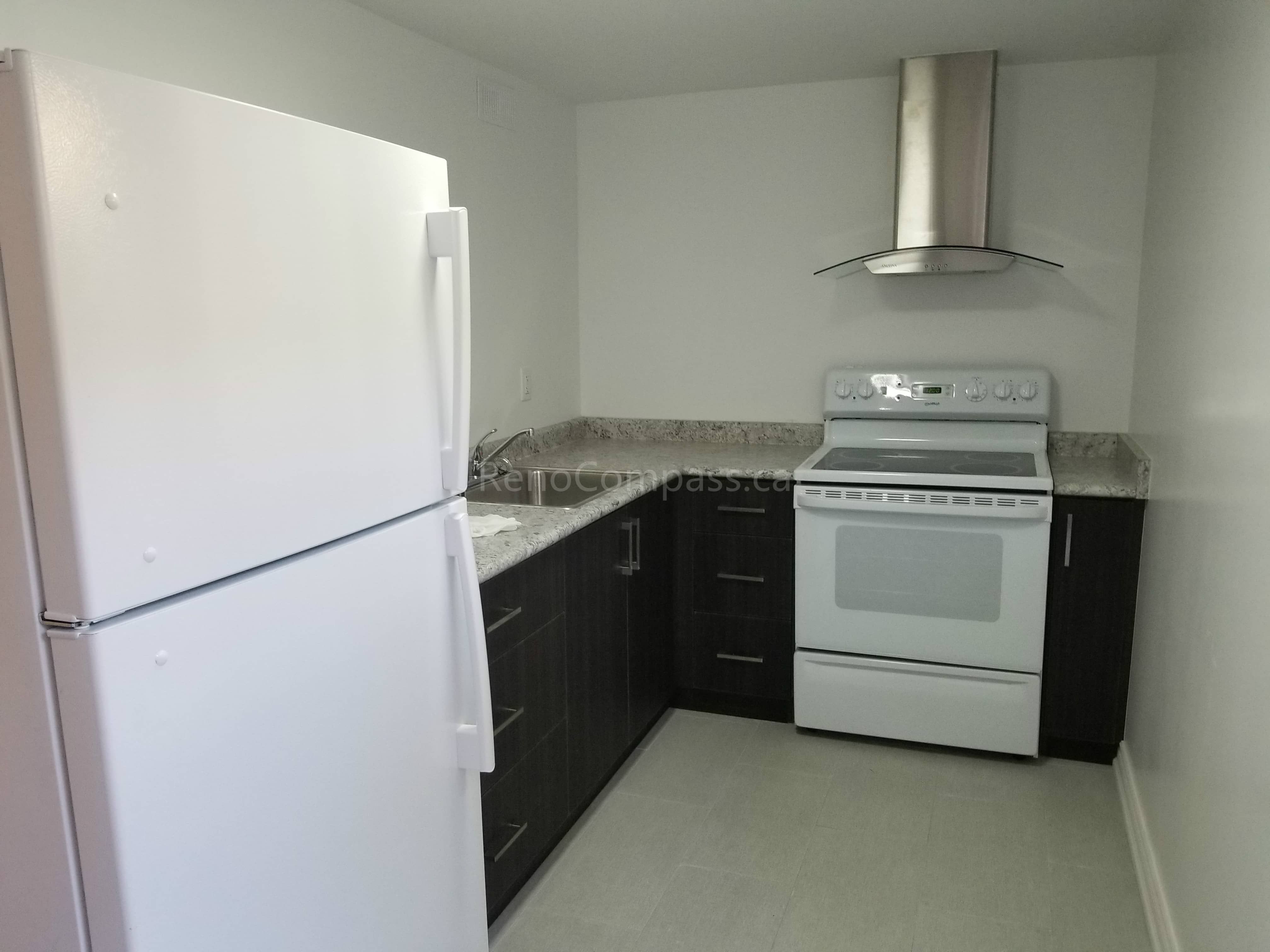 This Toronto basement renovation features a clean and functional modern kitchen designed for a basement apartment. The space includes dark wood cabinetry, quartz-style countertops, stainless steel range hood, full-size refrigerator, and durable flooring. Reno Compass Inc. optimized the layout to maximize storage and usability while maintaining a bright, minimalist look, creating a comfortable and practical basement living space for rental or family use.