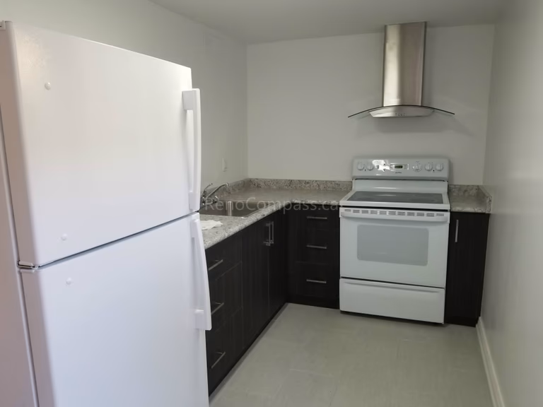 This Toronto basement renovation features a clean and functional modern kitchen designed for a basement apartment. The space includes dark wood cabinetry, quartz-style countertops, stainless steel range hood, full-size refrigerator, and durable flooring. Reno Compass Inc. optimized the layout to maximize storage and usability while maintaining a bright, minimalist look, creating a comfortable and practical basement living space for rental or family use.