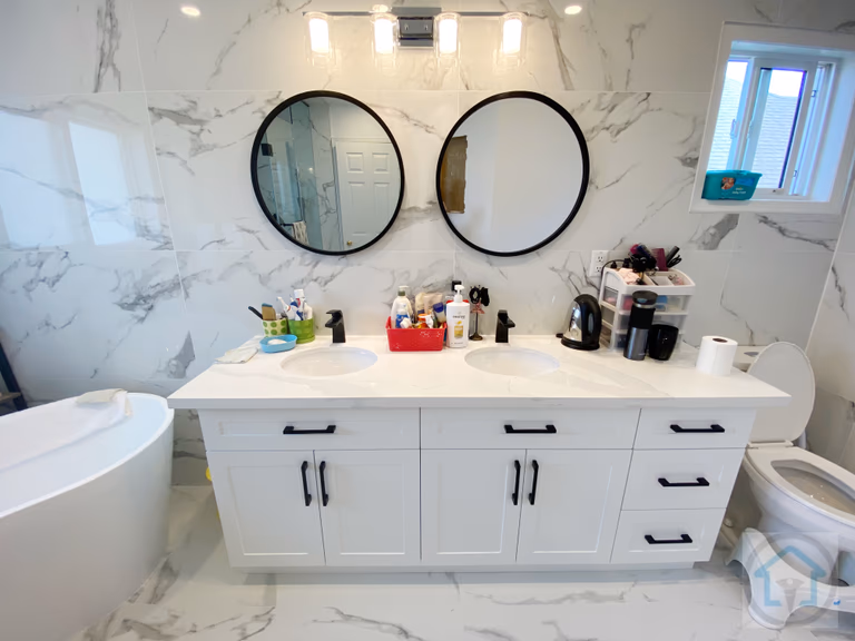 This Richmond Hill bathroom renovation features a custom double-sink vanity paired with round black-framed mirrors and modern wall-mounted lighting. The white shaker-style cabinetry is complemented by black hardware, creating a clean and balanced contrast against the marble-look wall tiles. Designed and completed by Reno Compass Inc., this bathroom remodel improves daily functionality while maintaining a timeless, easy-to-maintain aesthetic for Richmond Hill homes.