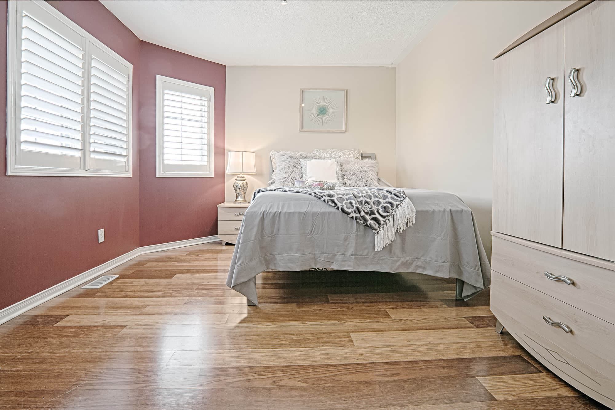 This cozy bedroom interior features a warm accent wall, natural hardwood flooring, and soft patterned bedding that create an inviting and comfortable atmosphere. The coordinated dresser set, arched mirror, and large window with plantation shutters enhance both functionality and natural light. A beautiful example of warm, minimalist bedroom interior design ideal for homeowners seeking stylish and comfortable home renovation inspiration in Toronto and the GTA.
