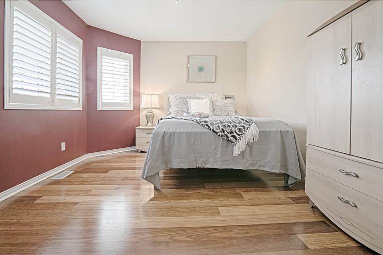 This cozy bedroom interior features a warm accent wall, natural hardwood flooring, and soft patterned bedding that create an inviting and comfortable atmosphere. The coordinated dresser set, arched mirror, and large window with plantation shutters enhance both functionality and natural light. A beautiful example of warm, minimalist bedroom interior design ideal for homeowners seeking stylish and comfortable home renovation inspiration in Toronto and the GTA.