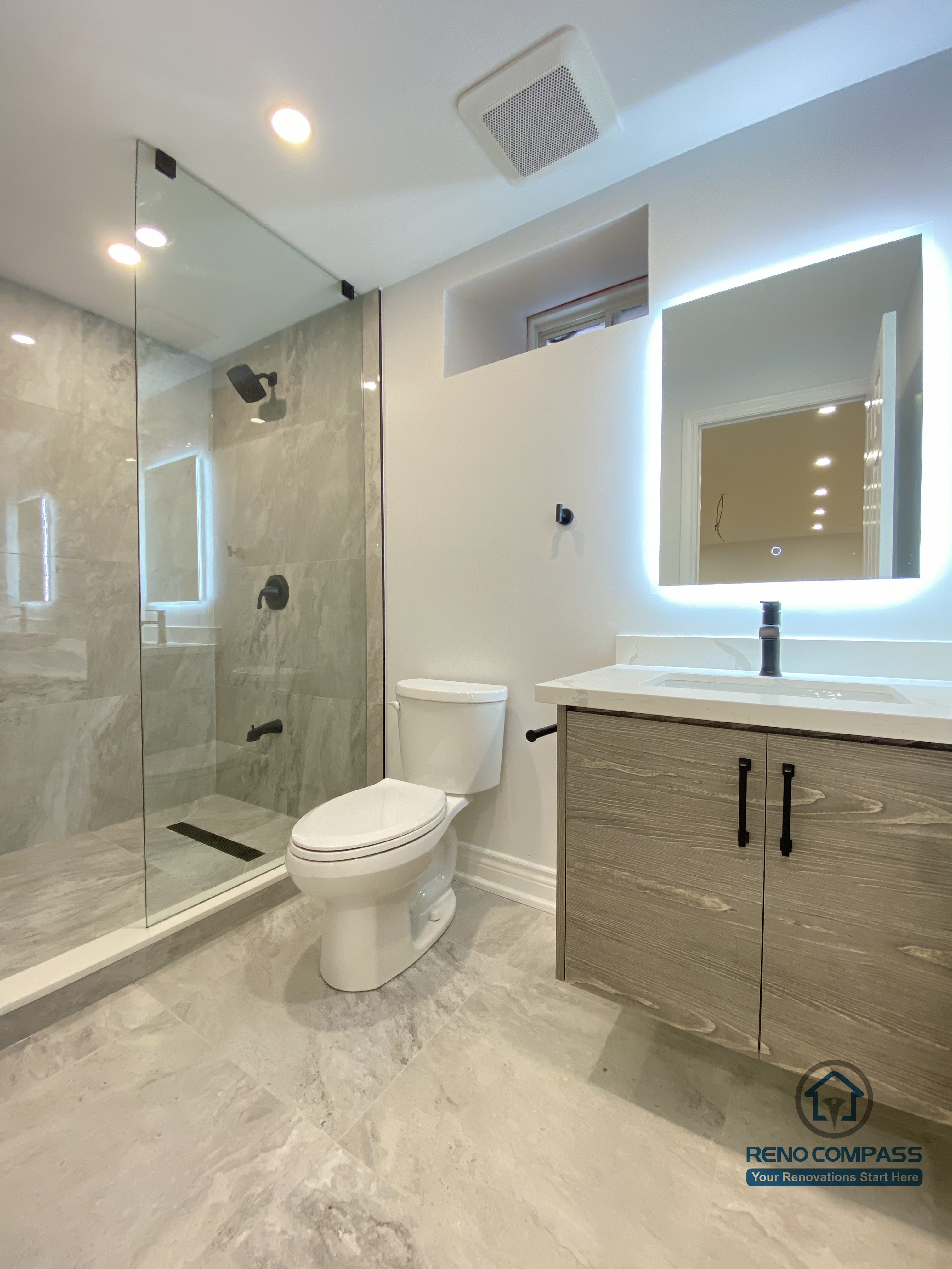This Markham bathroom renovation showcases a modern walk-in shower enclosed with frameless glass and finished in marble-look porcelain tile for a clean, seamless appearance. A floating vanity with wood-grain cabinetry adds warmth while maximizing floor space, paired with an LED backlit mirror to enhance lighting and daily functionality. Matte black fixtures and carefully planned ventilation improve both performance and durability. Completed by a trusted Toronto renovation company, this bathroom design focuses on comfort, waterproof detailing, and long-term residential value.