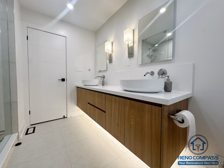 This Oakville bathroom renovation features a floating wood vanity paired with dual vessel sinks and a seamless quartz countertop. Integrated under-vanity lighting enhances the sense of space, while wall-mounted mirrors and modern sconces provide balanced, functional illumination. Clean-lined cabinetry and light porcelain flooring contribute to a refined, contemporary aesthetic designed for both daily use and long-term durability. Completed by Reno Compass, this bathroom reflects a modern approach to comfort and precision craftsmanship.