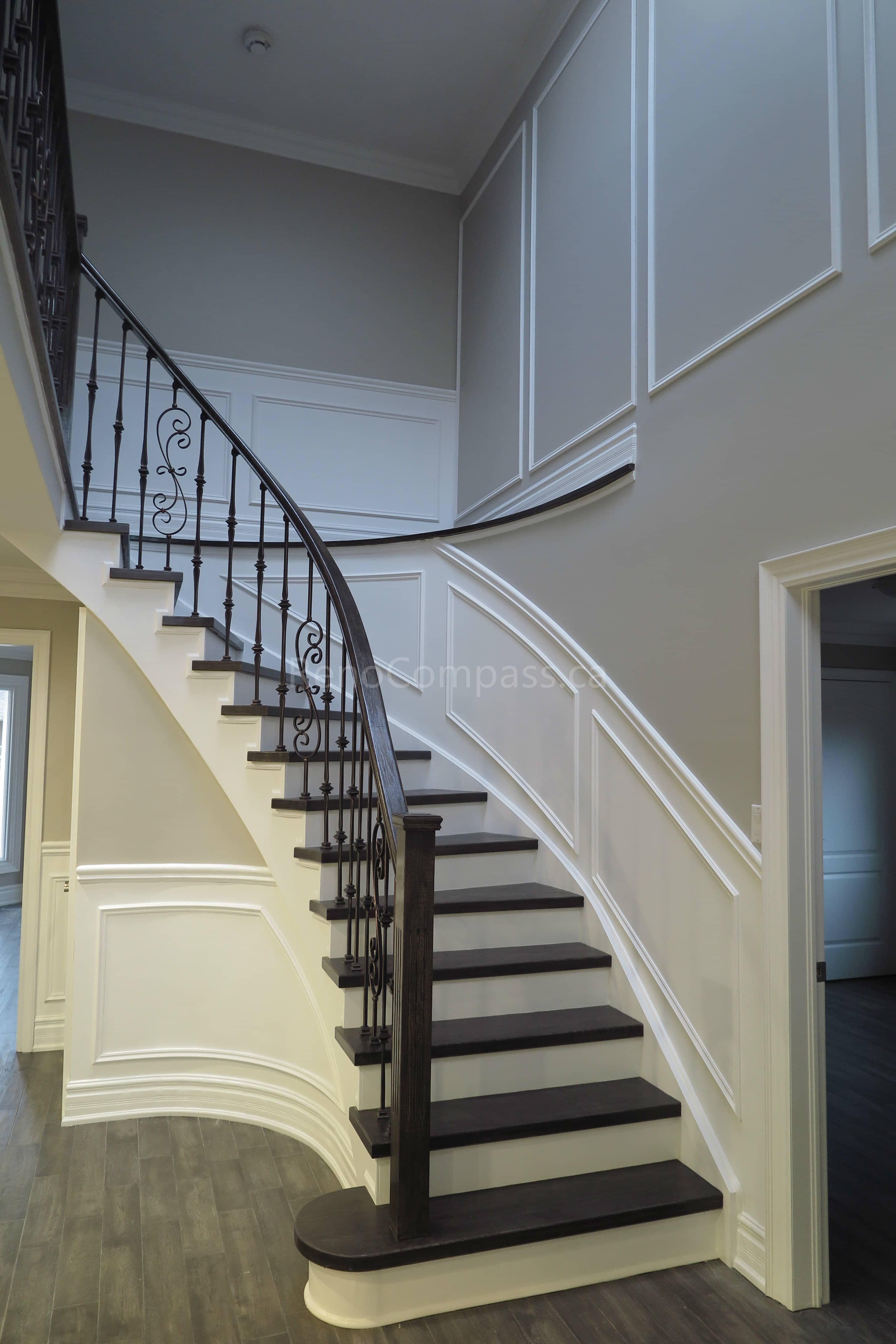 This Markham interior renovation showcases a custom curved staircase with dark wood treads, white risers, and elegant wrought iron railings. The surrounding space features detailed wall panel molding, soft neutral wall tones, and modern flooring that enhances the luxurious and timeless interior design. Completed by Reno Compass, a professional renovation contractor specializing in high-end interior renovations and custom staircase upgrades across Markham and the Greater Toronto Area.