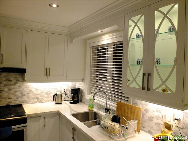 A bright and functional kitchen remodel in North York featuring a stunning white waterfall quartz island. This open-concept design seamlessly connects the cooking area to the dining space, highlighting custom white cabinetry, a built-in dishwasher, and large-format porcelain floor tiles for a clean, contemporary look.