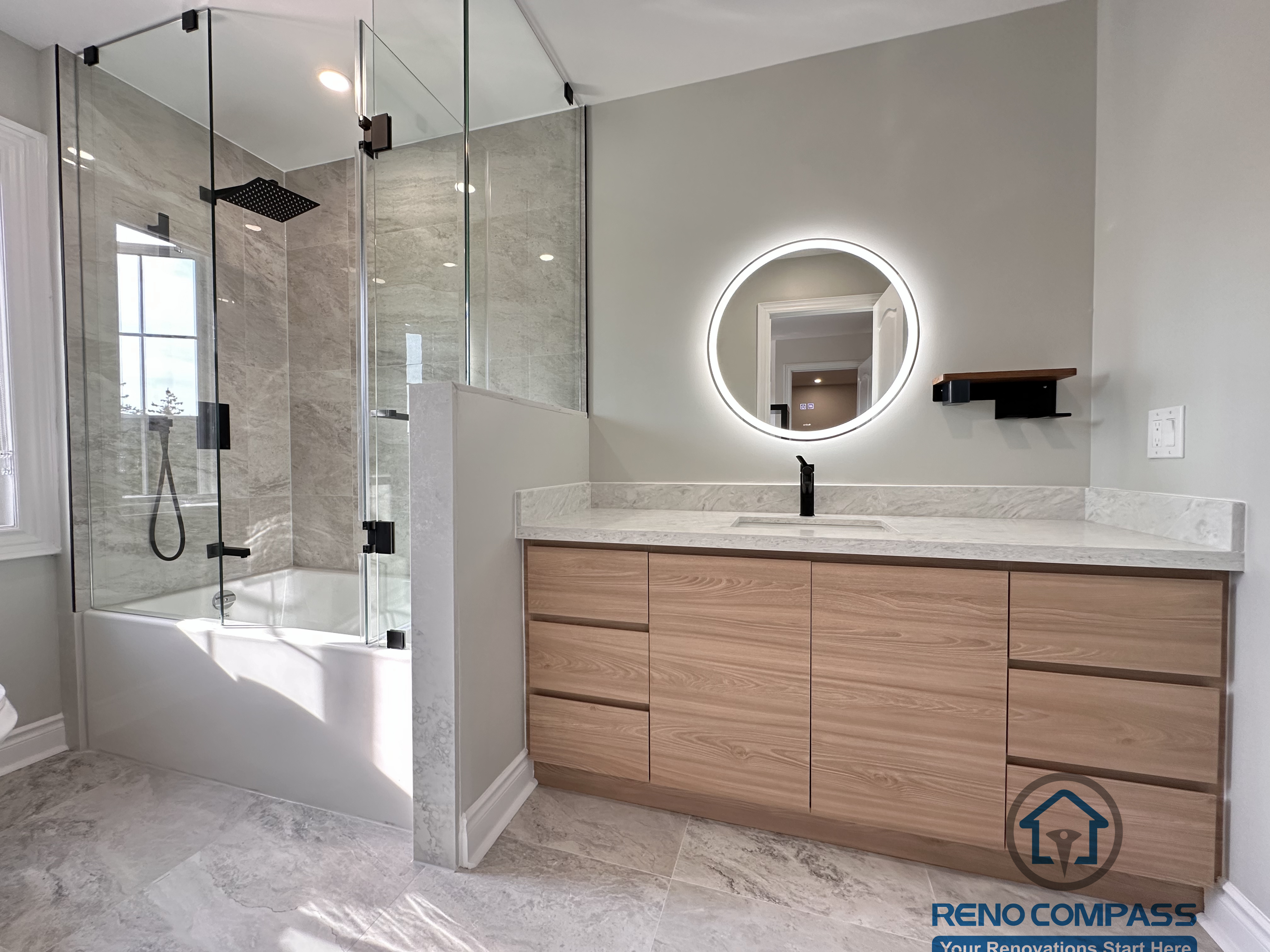 This Markham bathroom renovation features a custom wood-tone vanity with a quartz countertop, round LED backlit mirror, and matte black fixtures. The layout combines a glass-enclosed shower and bathtub with a practical vanity and toilet arrangement, creating a clean and well-balanced space. Designed and built by a professional Toronto renovation company, Reno Compass Inc., this bathroom emphasizes functionality, durability, and timeless design.