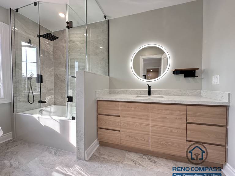 This Markham bathroom renovation features a custom wood-tone vanity with a quartz countertop, round LED backlit mirror, and matte black fixtures. The layout combines a glass-enclosed shower and bathtub with a practical vanity and toilet arrangement, creating a clean and well-balanced space. Designed and built by a professional Toronto renovation company, Reno Compass Inc., this bathroom emphasizes functionality, durability, and timeless design.