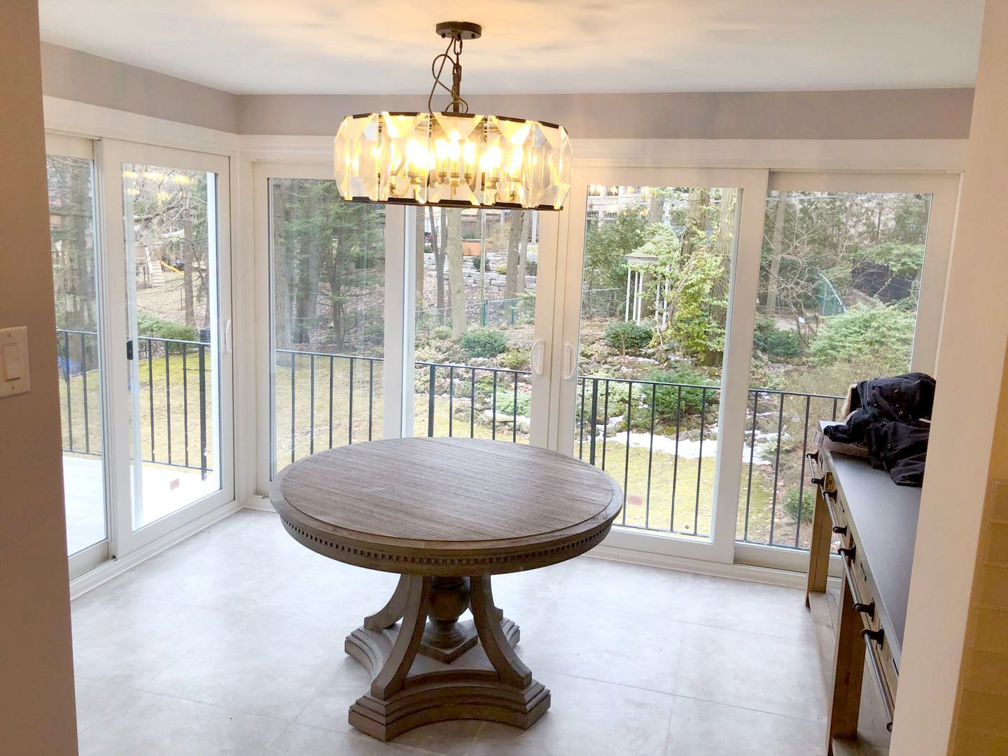 This Etobicoke interior renovation features a bright dining area surrounded by large windows and sliding glass doors, allowing abundant natural light and garden views. A modern chandelier adds a warm focal point above the dining space, while upgraded flooring and clean wall finishes create a refined and comfortable atmosphere. Designed and completed by Reno Compass Inc., this renovation enhances openness, natural light, and everyday functionality with a timeless interior design.