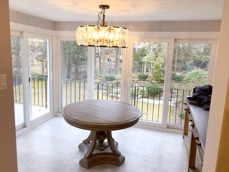 This Etobicoke interior renovation features a bright dining area surrounded by large windows and sliding glass doors, allowing abundant natural light and garden views. A modern chandelier adds a warm focal point above the dining space, while upgraded flooring and clean wall finishes create a refined and comfortable atmosphere. Designed and completed by Reno Compass Inc., this renovation enhances openness, natural light, and everyday functionality with a timeless interior design.