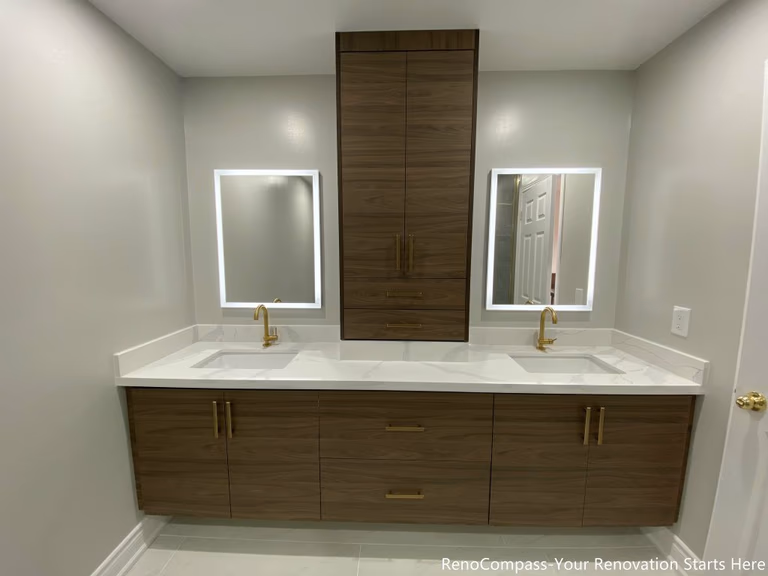 This Oakville master bathroom renovation showcases a modern double-sink vanity with custom wood cabinetry, quartz countertop, and elegant gold fixtures. The design features large-format floor tiles, LED backlit mirrors, and a sleek glass shower, creating a bright and luxurious spa-like atmosphere. Completed by a professional Toronto renovation company, this bathroom upgrade focuses on functionality, storage, and contemporary style, enhancing both comfort and property value for the homeowner.