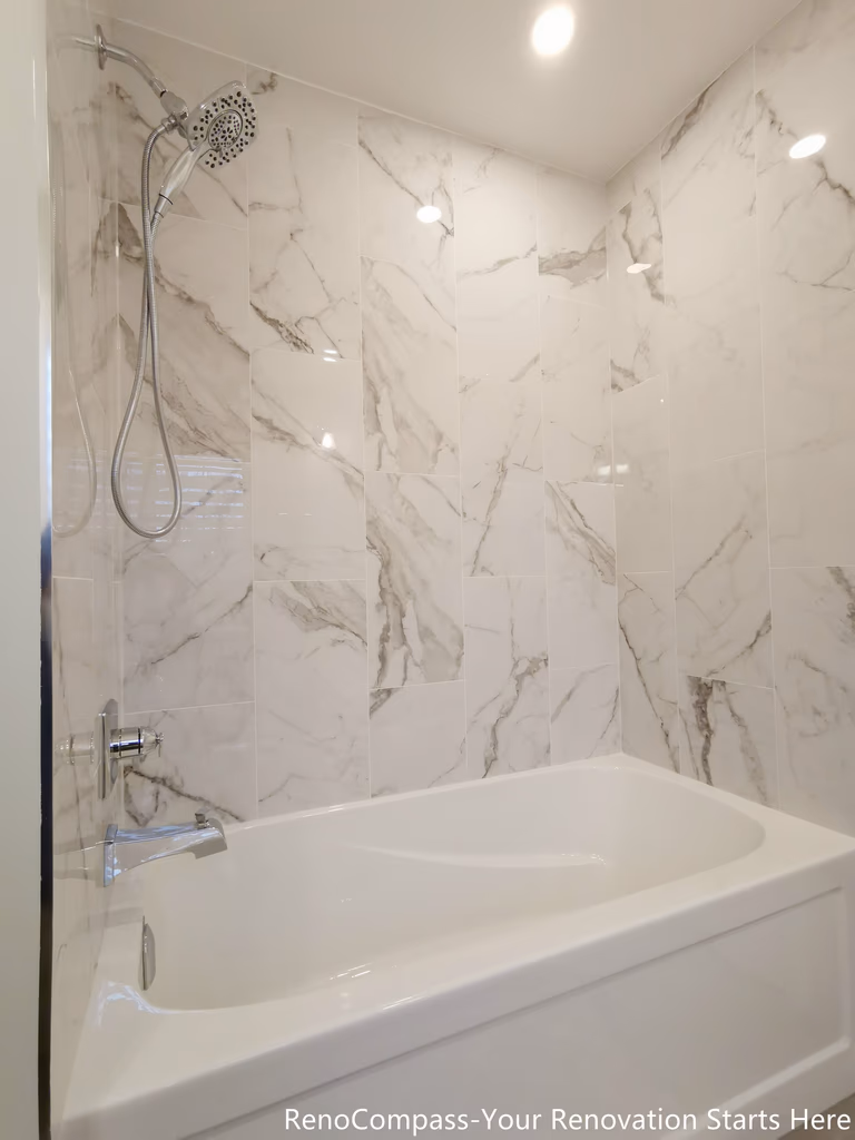 This image highlights the bathtub and shower area in an Oakville bathroom renovation, featuring full-height marble-look wall panels that create a clean and seamless backdrop. The integrated bathtub is paired with a wall-mounted shower system, balancing everyday practicality with a spa-inspired aesthetic. Recessed ceiling lighting enhances the natural veining of the stone surfaces, giving the space a bright, refined, and timeless finish.