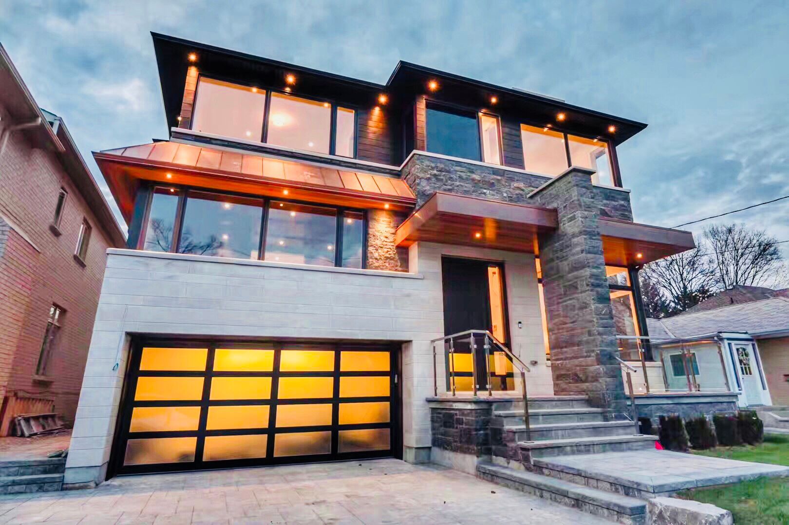 An architectural masterpiece in Toronto featuring a blend of natural fieldstone, limestone, and premium copper roofing accents. This modern custom build showcases expansive floor-to-ceiling windows, a glass-panel garage door, and sophisticated soffit lighting for maximum evening curb appeal.