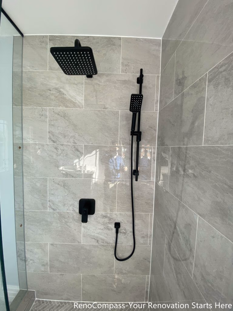 This Thornhill bathroom renovation features a modern walk-in shower enclosed with frameless glass panels, creating a bright and visually open space. The shower is finished with large-format gray wall tiles, a patterned shower base, and matte black fixtures for a clean, contemporary contrast. Natural light from the window enhances the overall brightness, while the glass enclosure replaces traditional drywall to significantly improve spatial flow, functionality, and long-term usability.