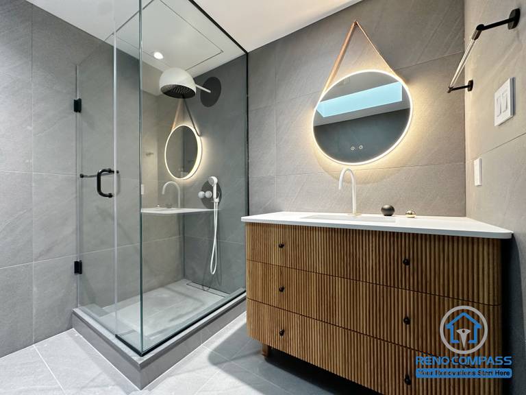 This overall view showcases a modern bathroom renovation in Richmond Hill, featuring a frameless glass shower, large-format grey tiles, and a custom wood-textured vanity with an integrated sink. Sculptural backlit mirrors and minimalist black fixtures add warmth and contrast, creating a balanced, spa-inspired atmosphere. Designed and completed by Reno Compass, this project reflects a refined contemporary bathroom renovation in the Greater Toronto Area.