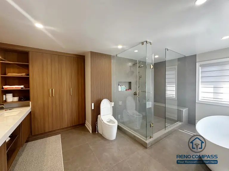 This image showcases a modern bathroom renovation completed in North York, featuring a spacious walk-in glass shower, custom wood vanity cabinetry, and built-in storage solutions. Large-format floor tiles enhance durability and visual continuity, while the frameless glass enclosure keeps the space open and bright. The combination of warm wood tones and neutral finishes creates a balanced, contemporary bathroom design focused on comfort, functionality, and everyday usability in North York homes.